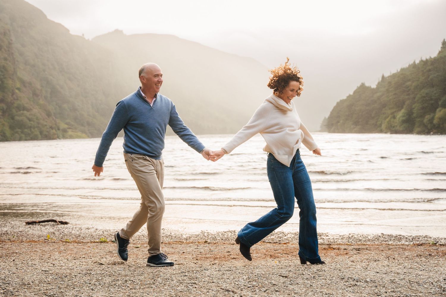 Two people hold hands while playfully walking along a misty beach with mountains in the background. couple's photographer dublin ireland