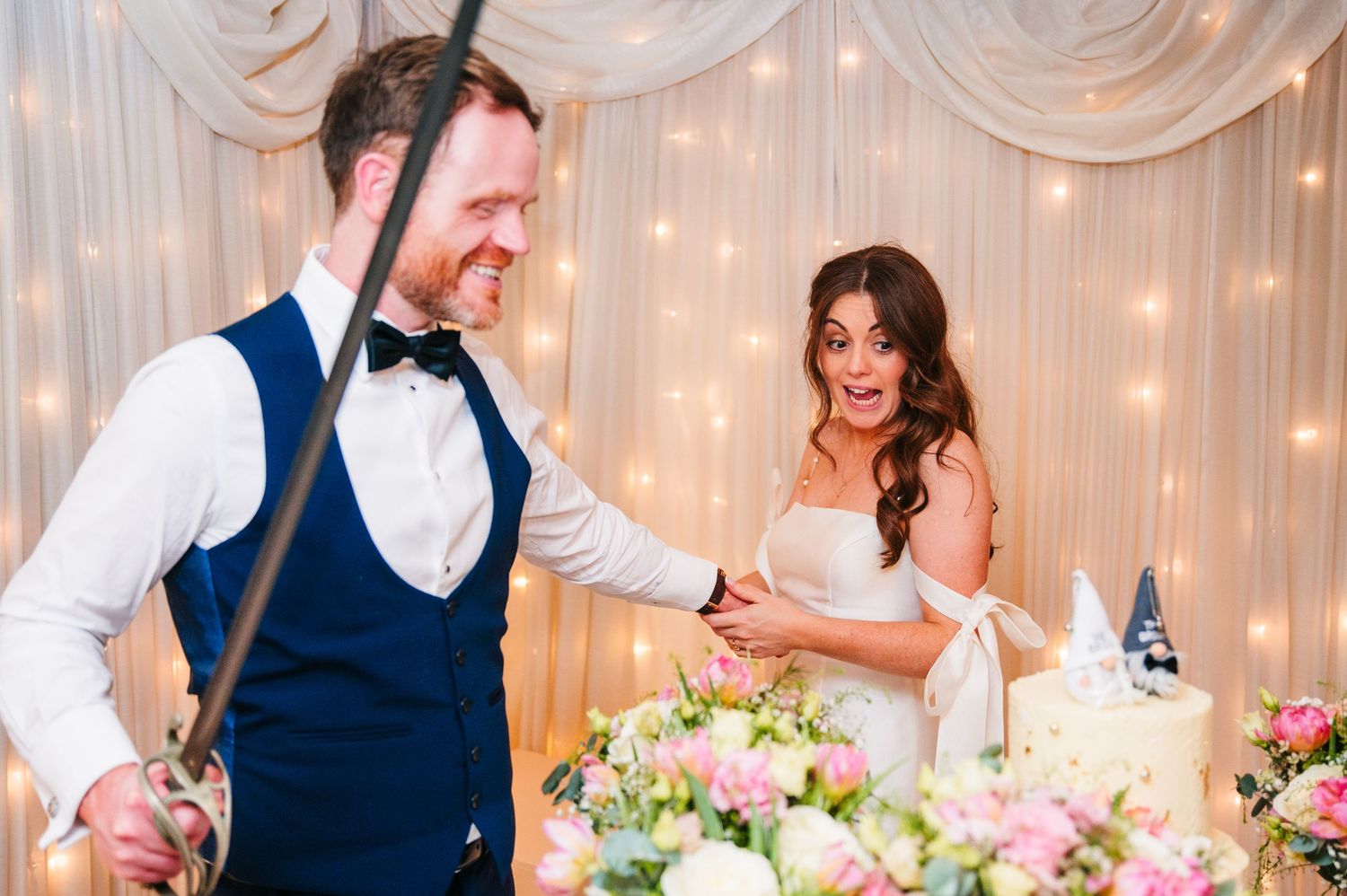 Newlyweds participate in their wedding reception cake cutting tradition with floral decor and twinkling lights. Wedding PHOTOGRAPHER Dublin
