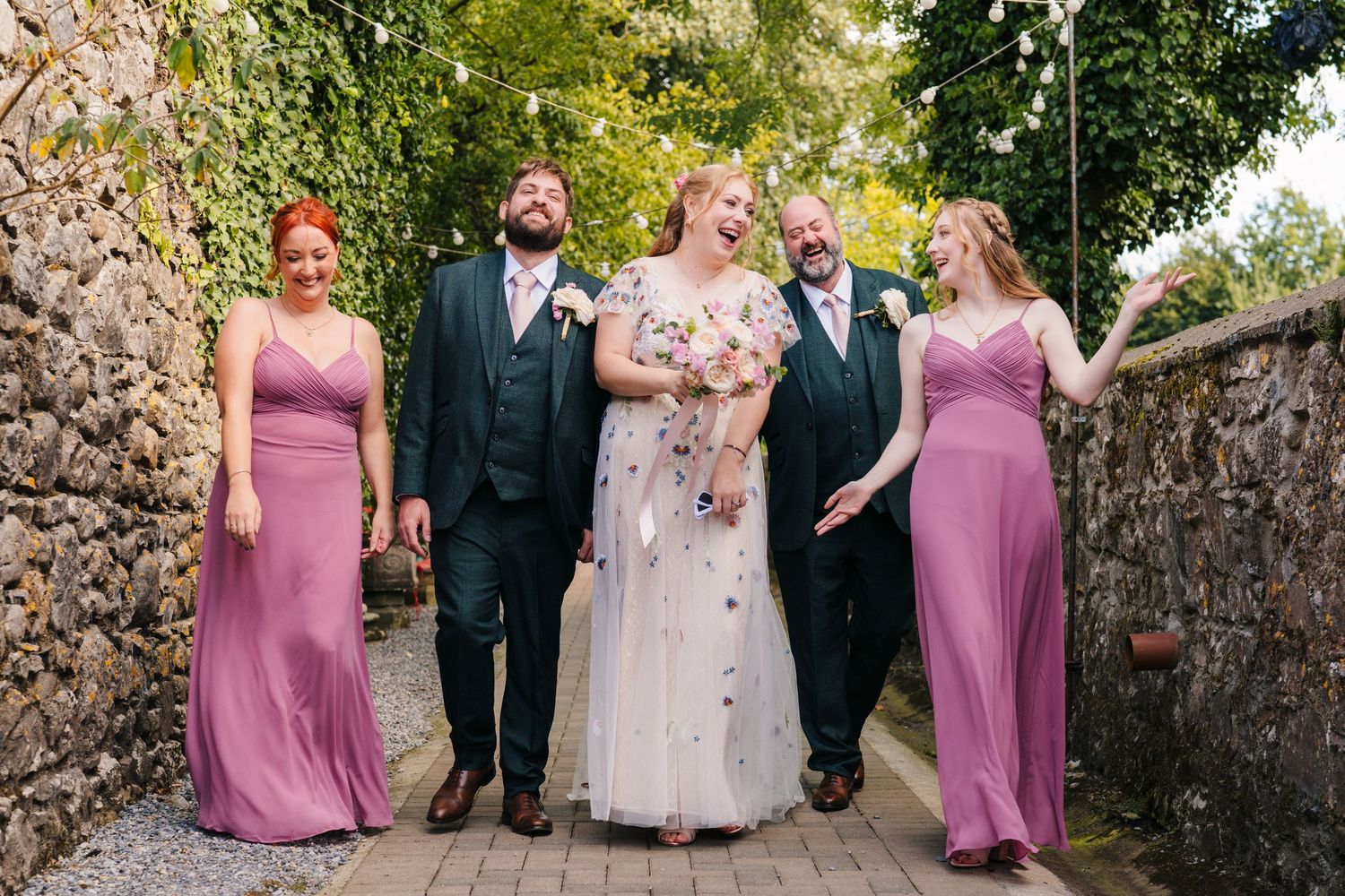 Wedding party in formal attire standing against stone wall with pink bridesmaids dresses creating an elegant scene. . Village Barn Tyrrellsp