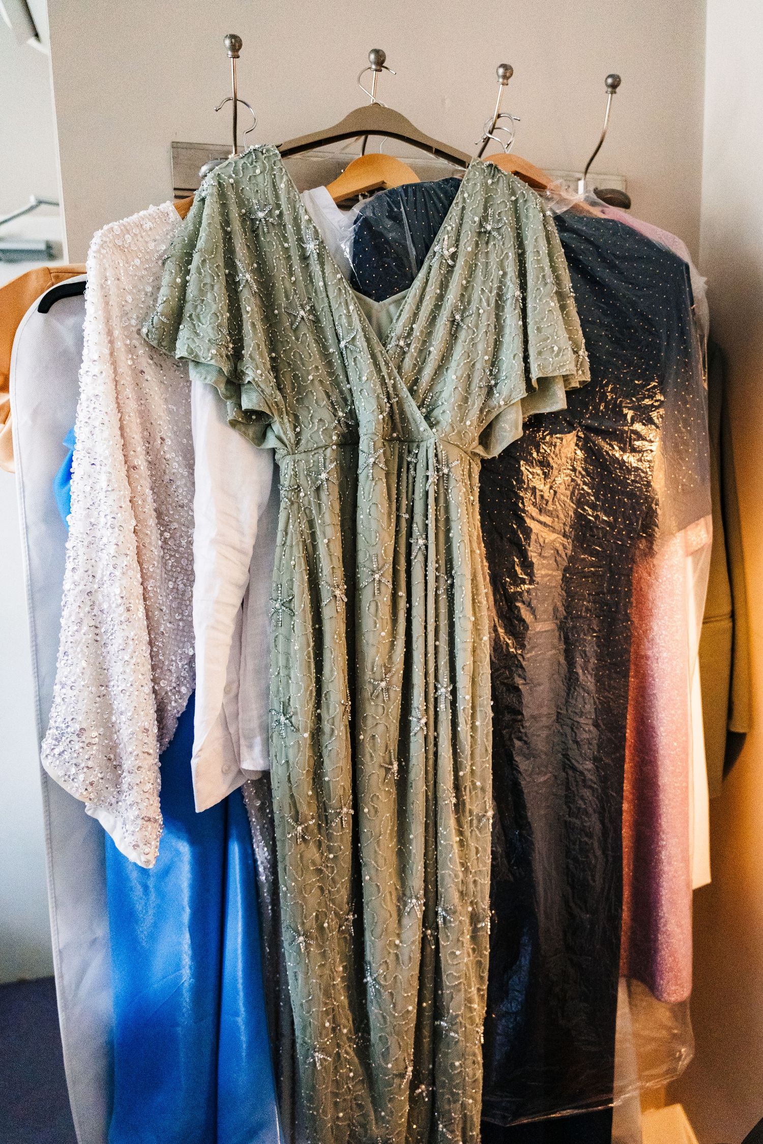 A sage green sequined dress with flutter sleeves hangs among other formalwear on clothing hooks. House Dublin Wedding Photographer