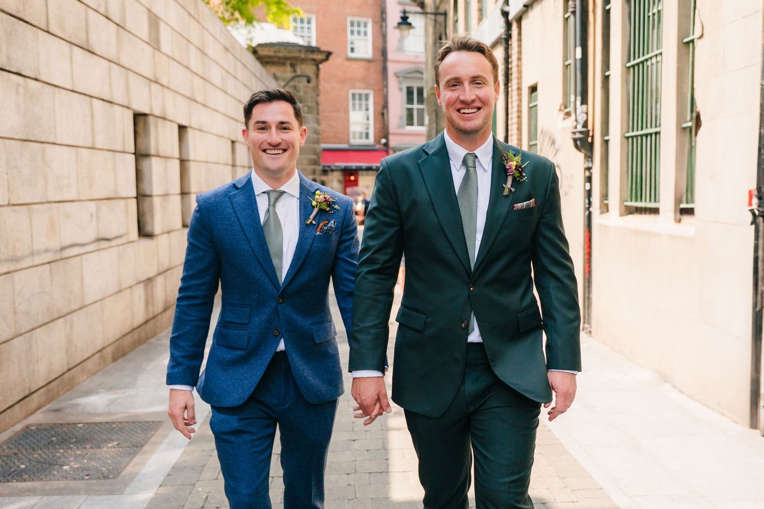 Two people in contrasting blue and green suits walking together along a bright urban street. Same sex wedding PHOTOGRAPHER Dublin