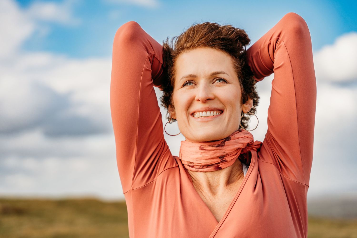 Person in a salmon-colored top stretches happily outdoors against a bright blue sky with clouds. Portrait PHOTOGRAPHER Dublin