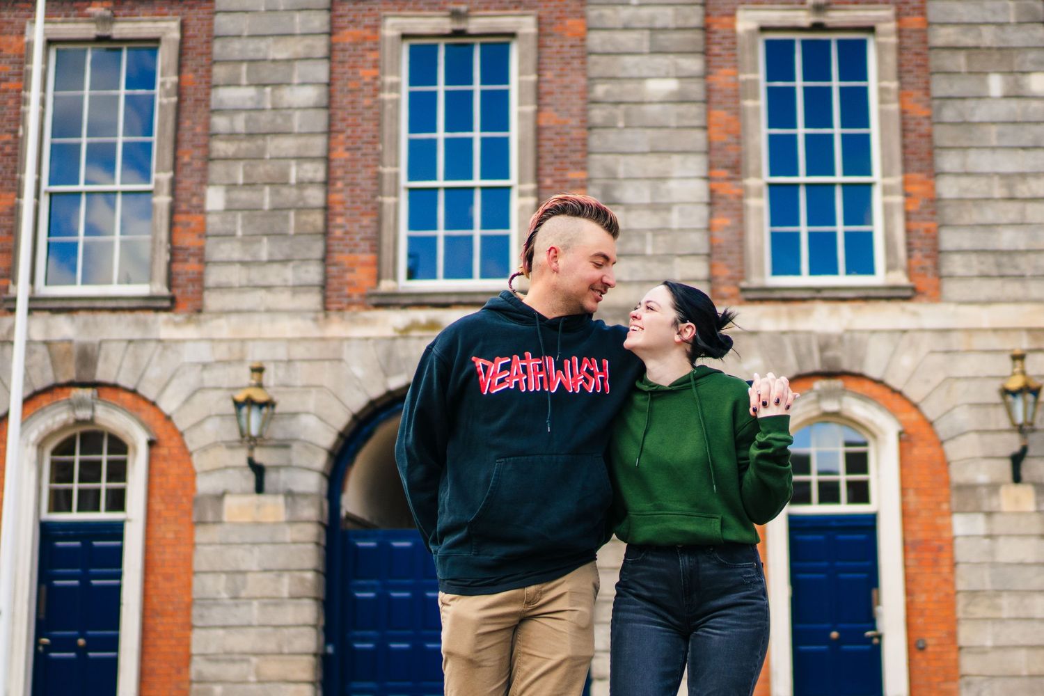 A couple shares a joyful moment in front of a historic brick building with blue doors. PHOTOGRAPHER Dublin Ireland