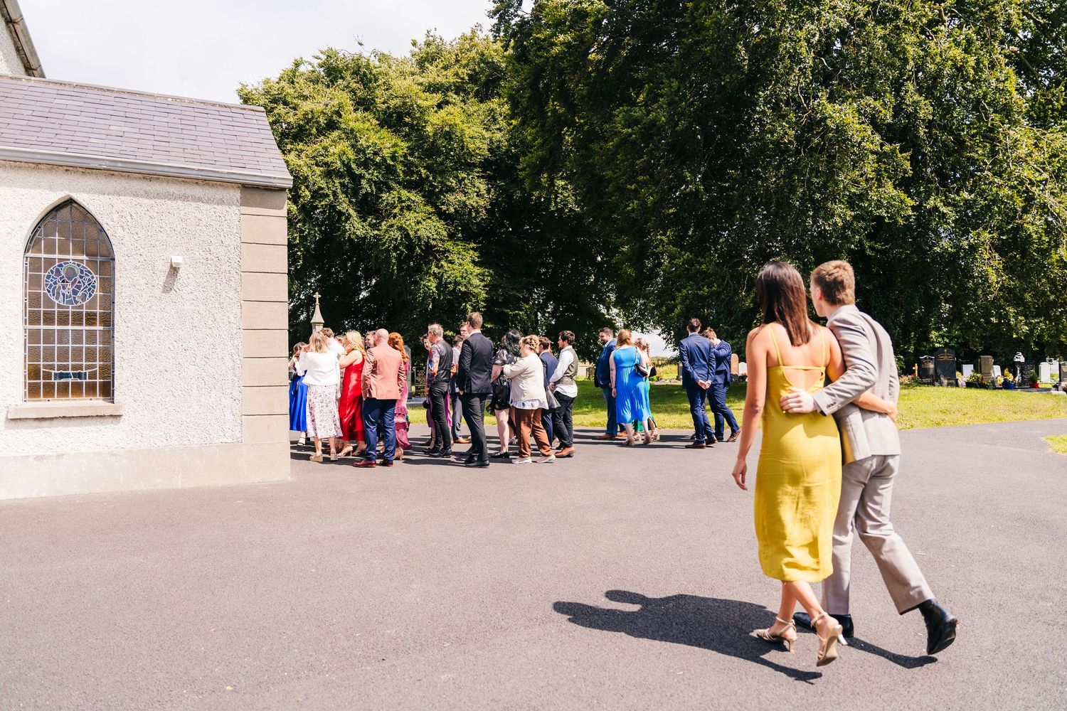 Millhouse slane wedding photographer dublin Wedding guests gathered outside a chapel with a couple in yellow dress and suit walking nearby.