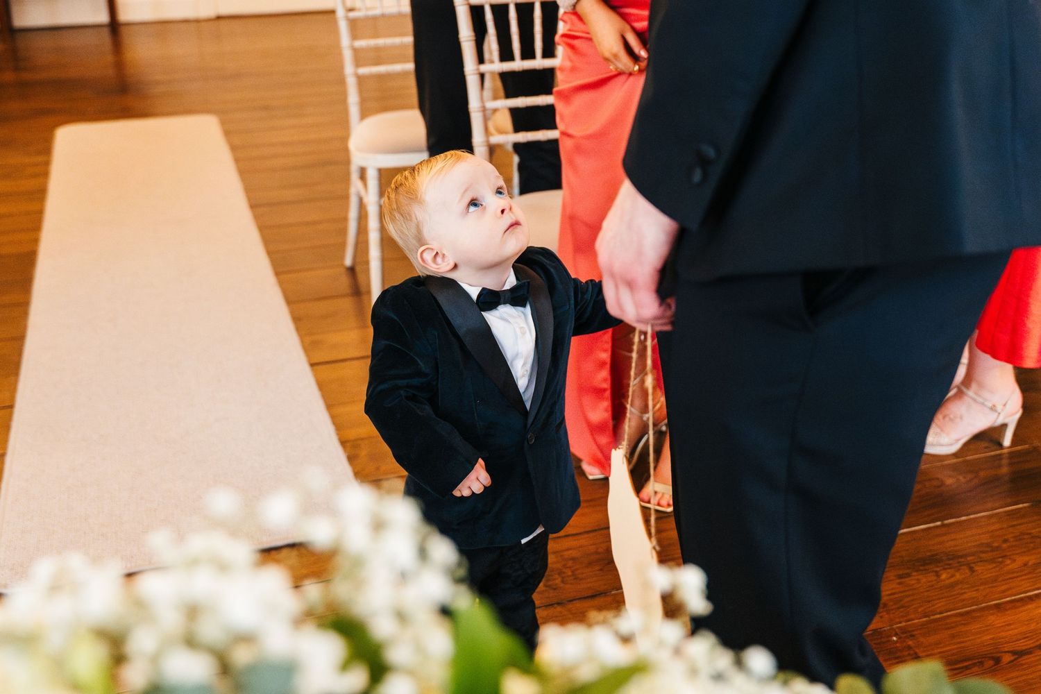 A small child in a black tuxedo and bowtie interacts with adults at a formal event on a hardwood floor.  Wedding PHOTOGRAPHER Dublin