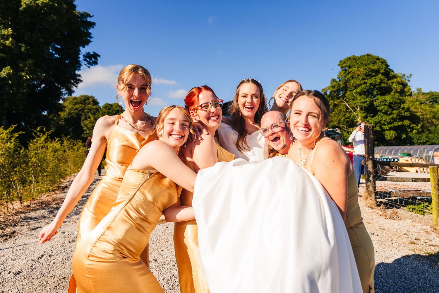 Poulaphouca Wedding photographer wicklow Joyful bridal party in gold dresses huddles together for fun outdoor photo on sunny day.