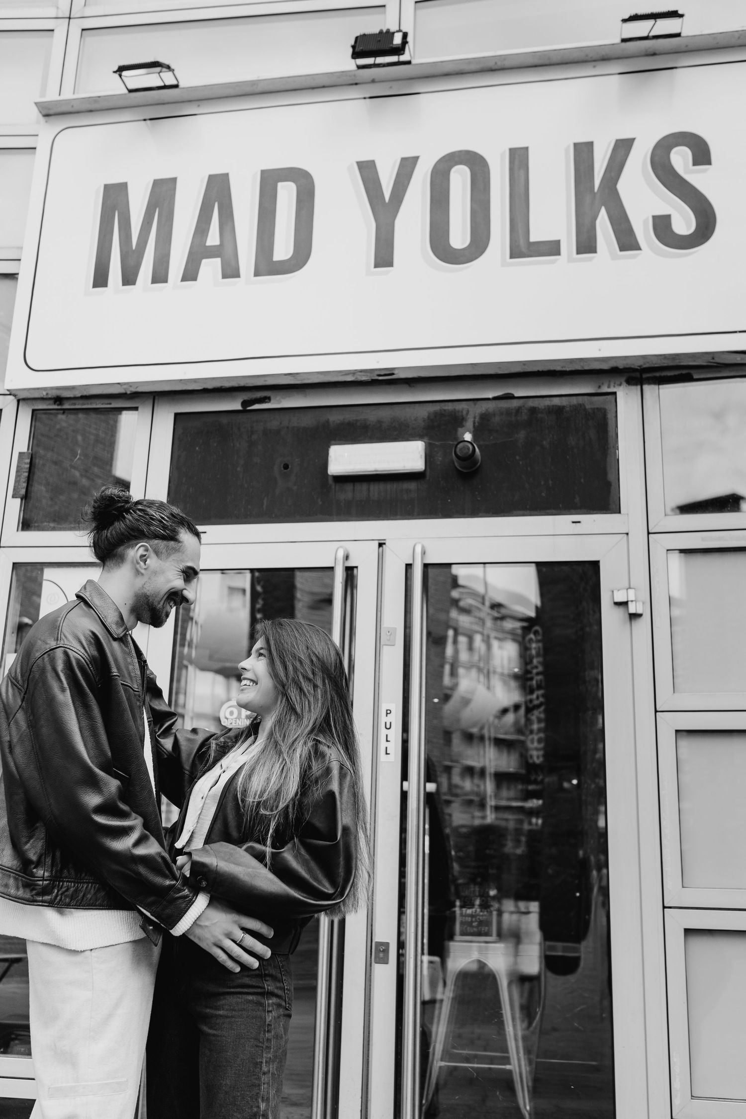 Black and white storefront photo showing a couple embracing outside a business called Mad Yolks.