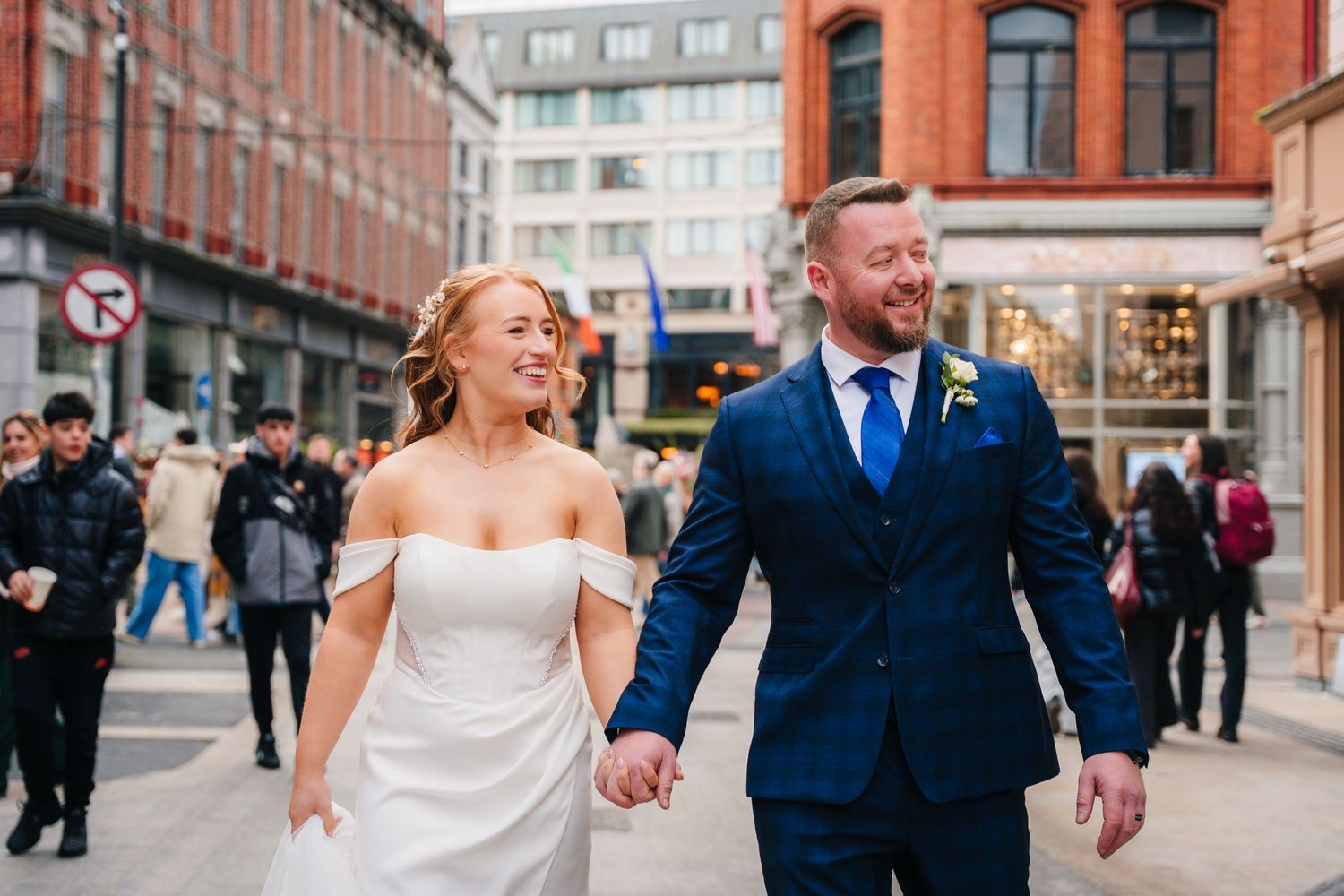 Newlywed couple walks through city street holding hands with brick buildings and urban architecture in background. Dublin wedding photogrAPh