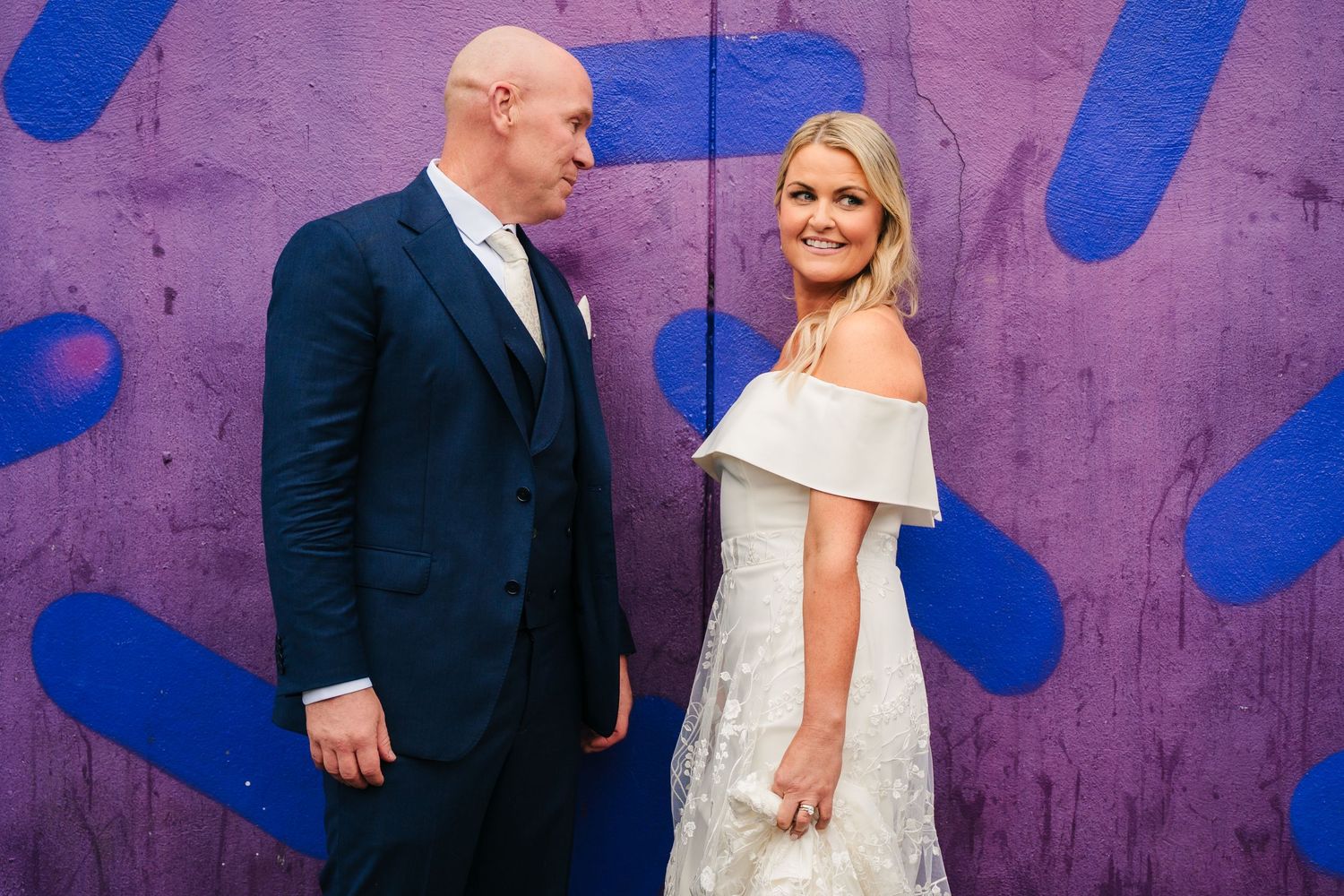 Couple poses against a purple wall with blue artistic strokes during their wedding photoshoot. The Dean Dublin Wedding Photographer