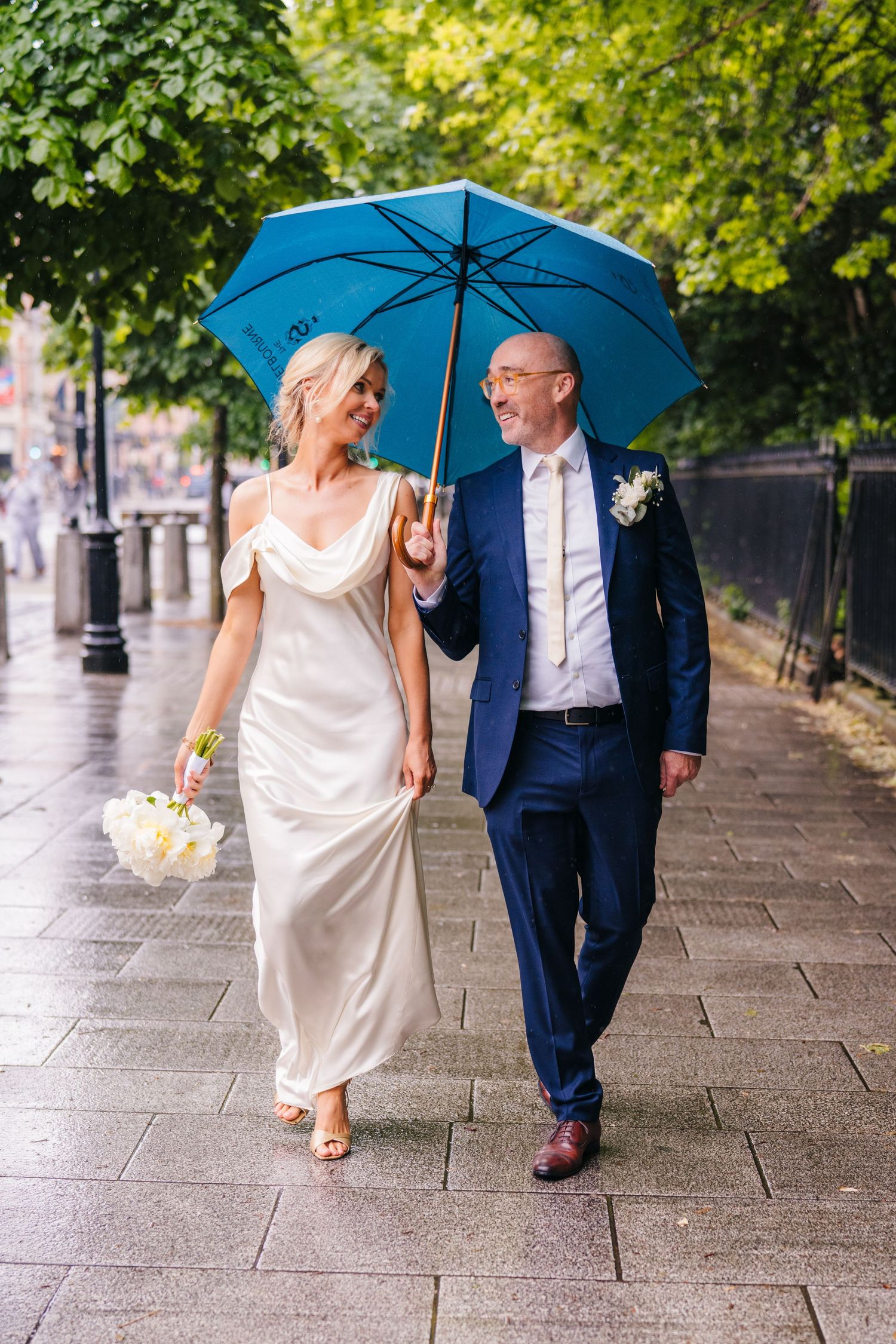 A couple shares a bright blue umbrella while walking together on a rainy cobblestone path in their wedding attire. Wedding PHOTOGRAPHER Dubl