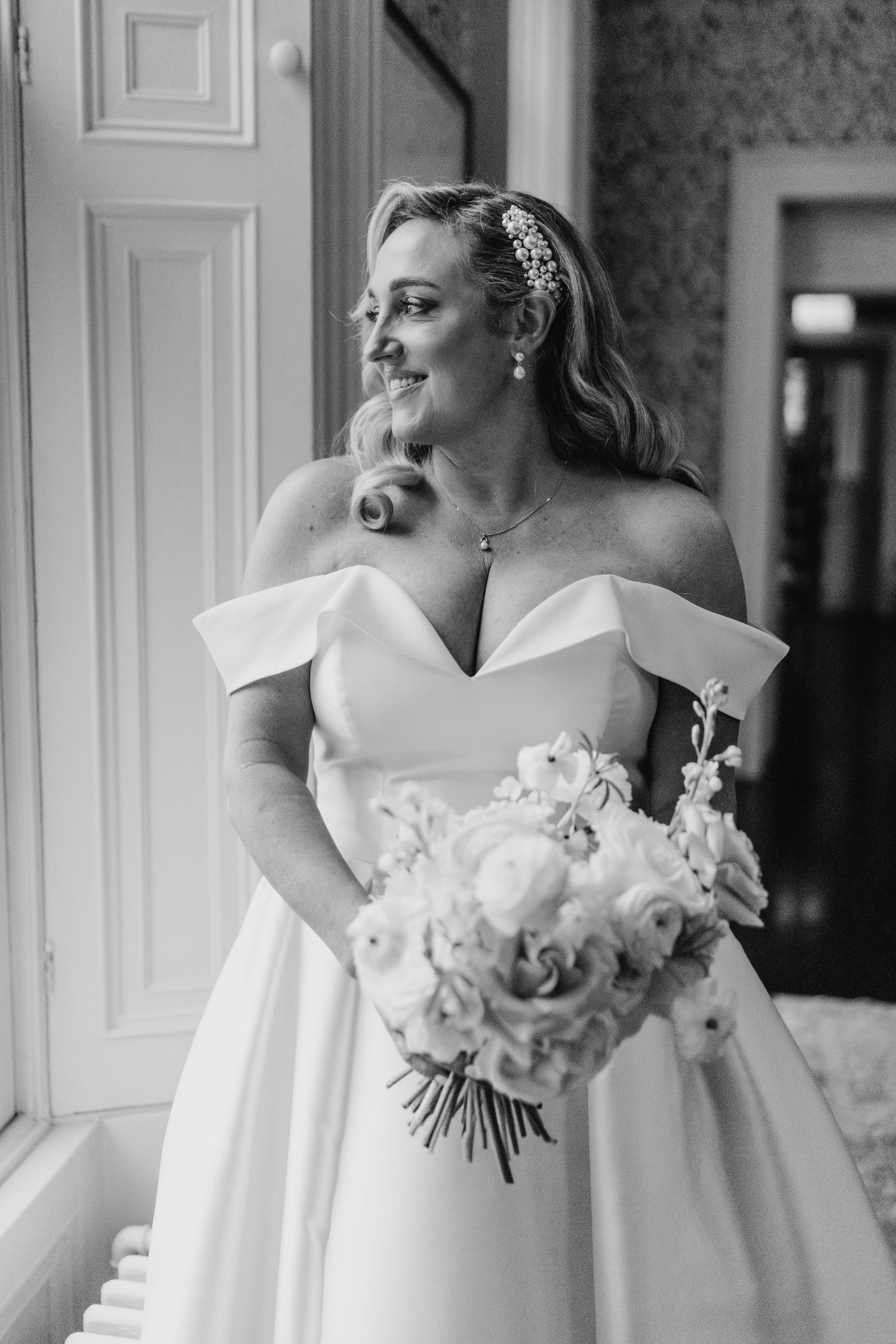 A bride in an elegant off-shoulder white wedding gown holds a beautiful bouquet of roses while smiling in black and white.