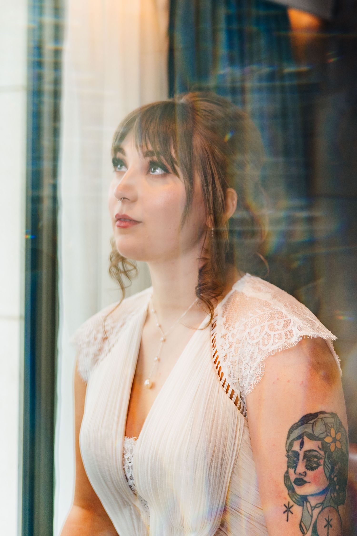 A person in a white lace dress with an arm tattoo gazes out of a window with soft lighting and reflections.