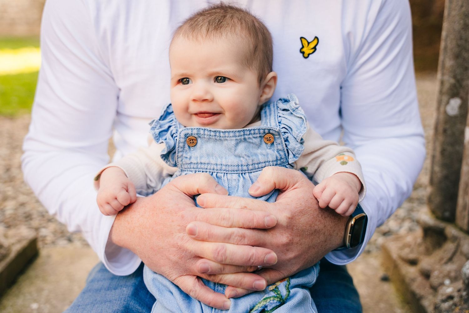A small baby wearing denim overalls sits securely in a parent's lap wearing a white shirt with yellow butterfly emblem. family PHOTOGRAPHER