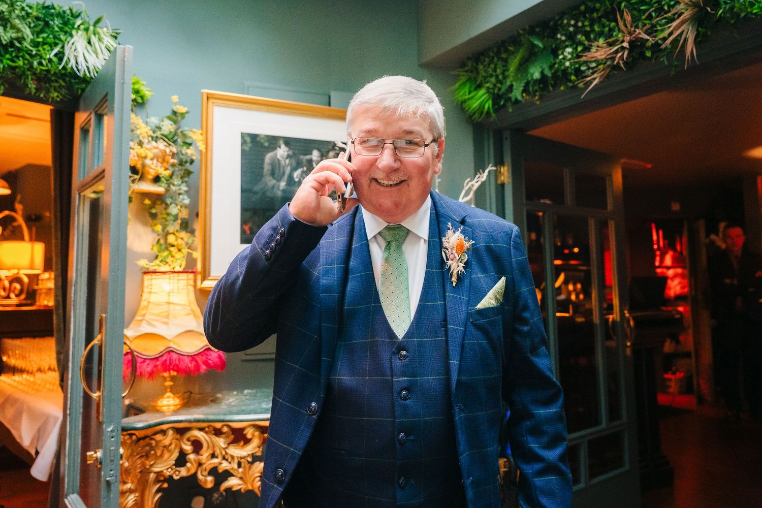 A gentleman in a blue three-piece suit poses elegantly in front of decorative indoor plants.