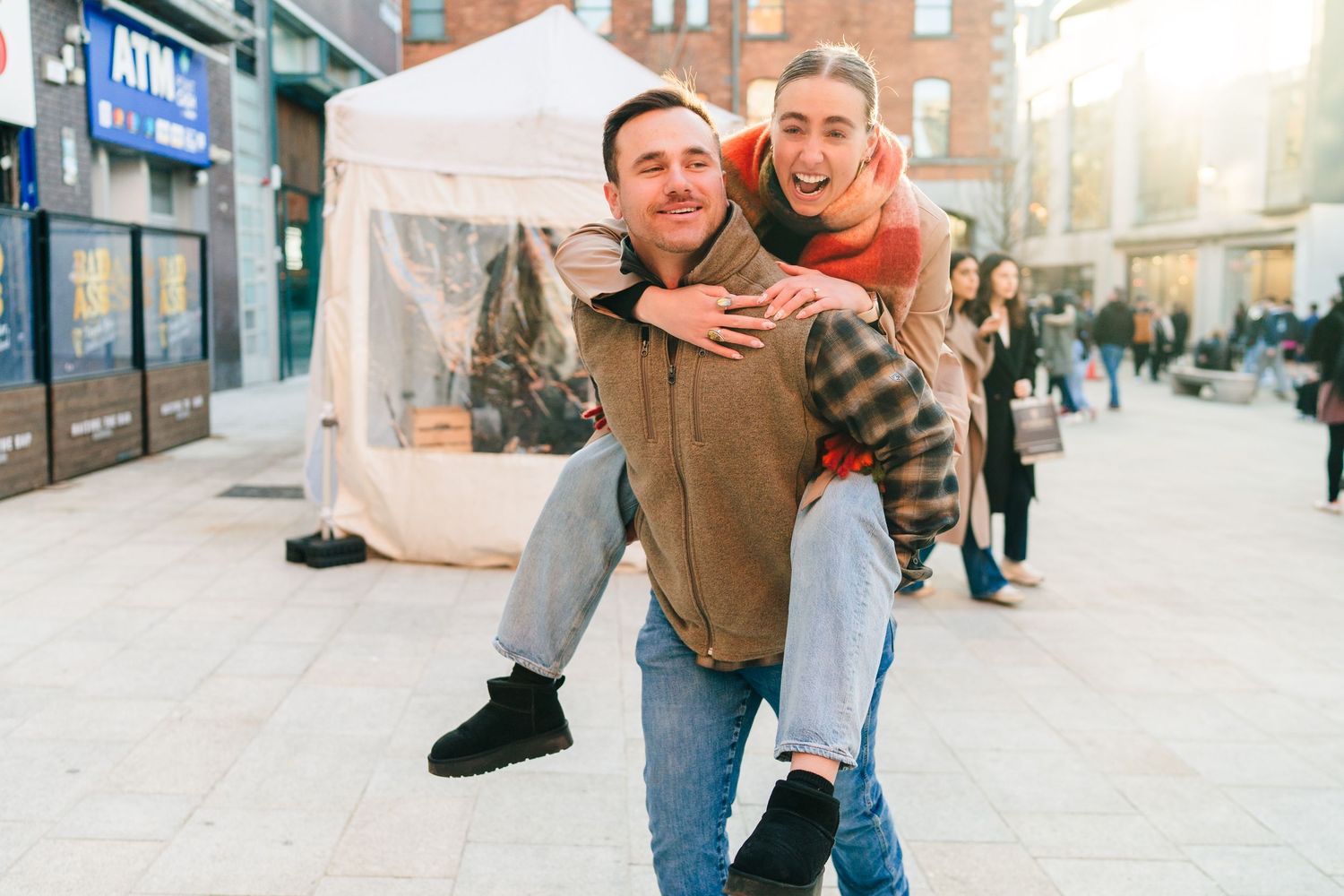 Two people enjoying a playful piggyback ride on a sunny urban street with storefronts in the background. PHOTOGRAPHER Dublin ireland