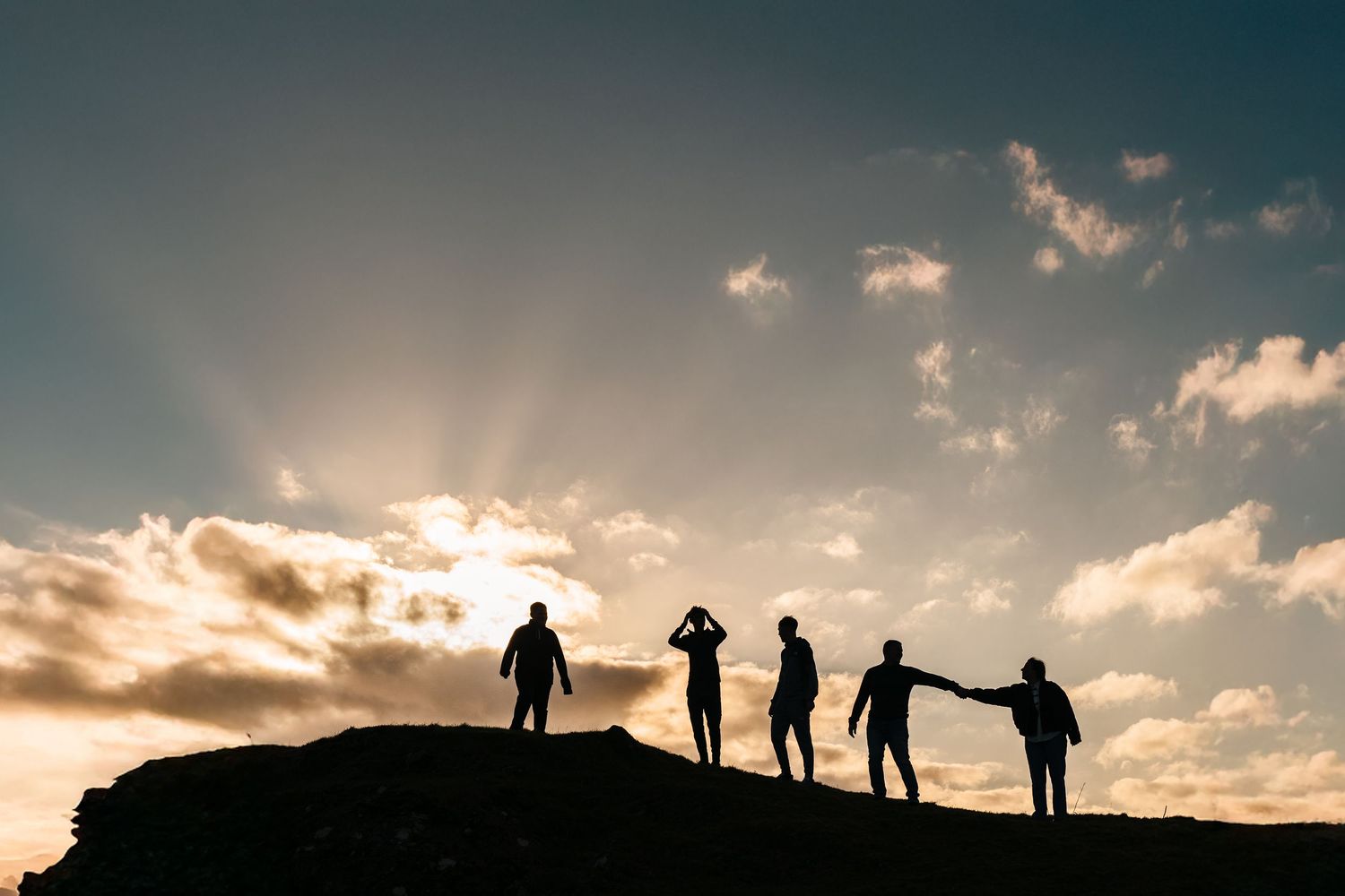 Silhouettes of people standing on hilltop against dramatic sunset sky with sunburst and clouds.