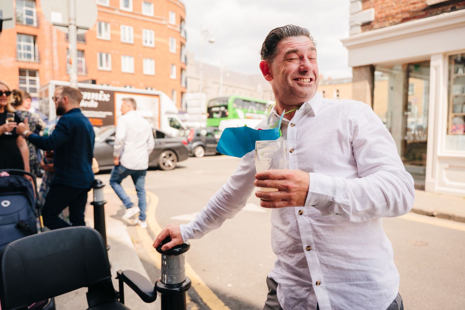 A joyful man in a white shirt laughs while holding drinks on a city street. Wedding Photographer Dublin Ireland