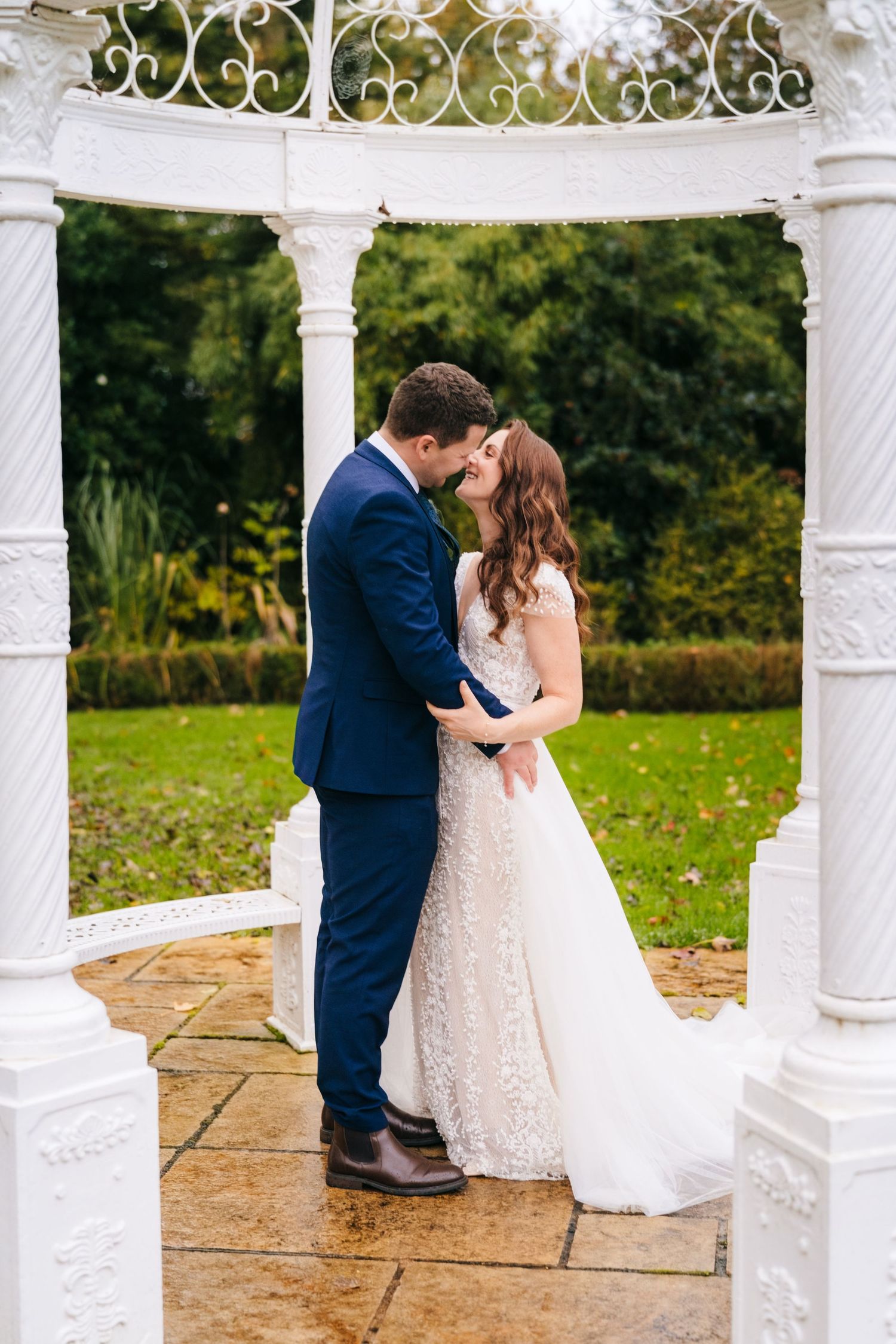 A romantic wedding couple shares a tender kiss under an elegant white gazebo on a stone patio surrounded by autumn foliage.