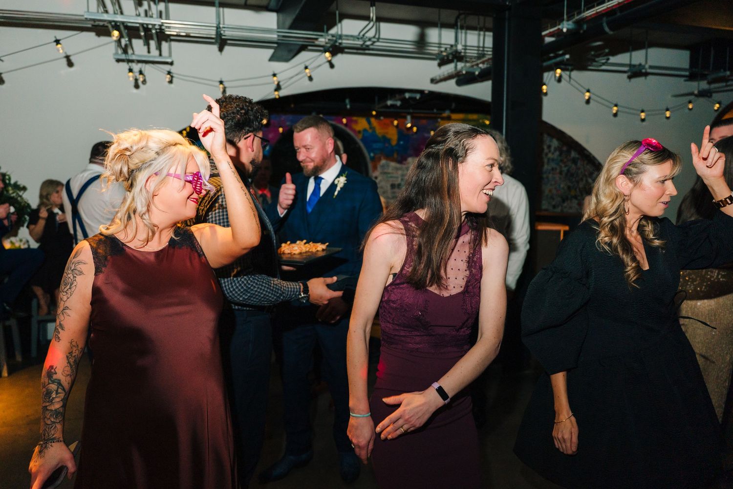 Wedding guests dancing and celebrating at an indoor reception with industrial-style lighting. Dublin wedding photogrAPher