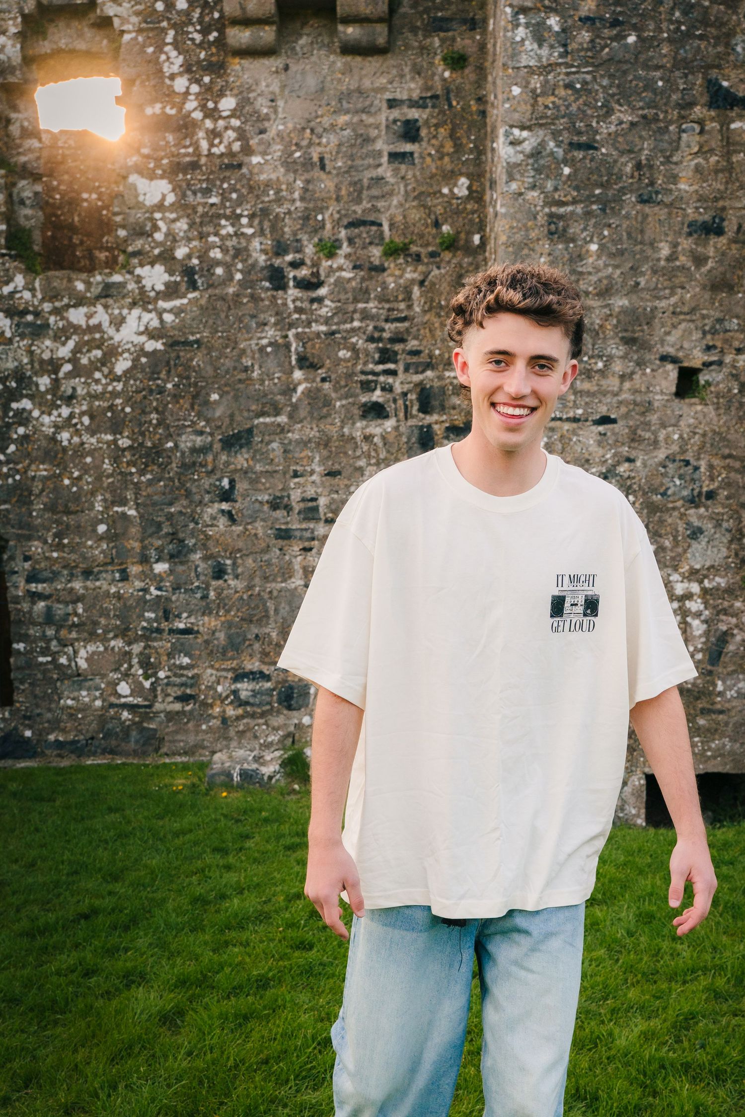 Person in a white t-shirt poses playfully against a rustic stone wall with green grass in the foreground during sunset. Photographer Ireland