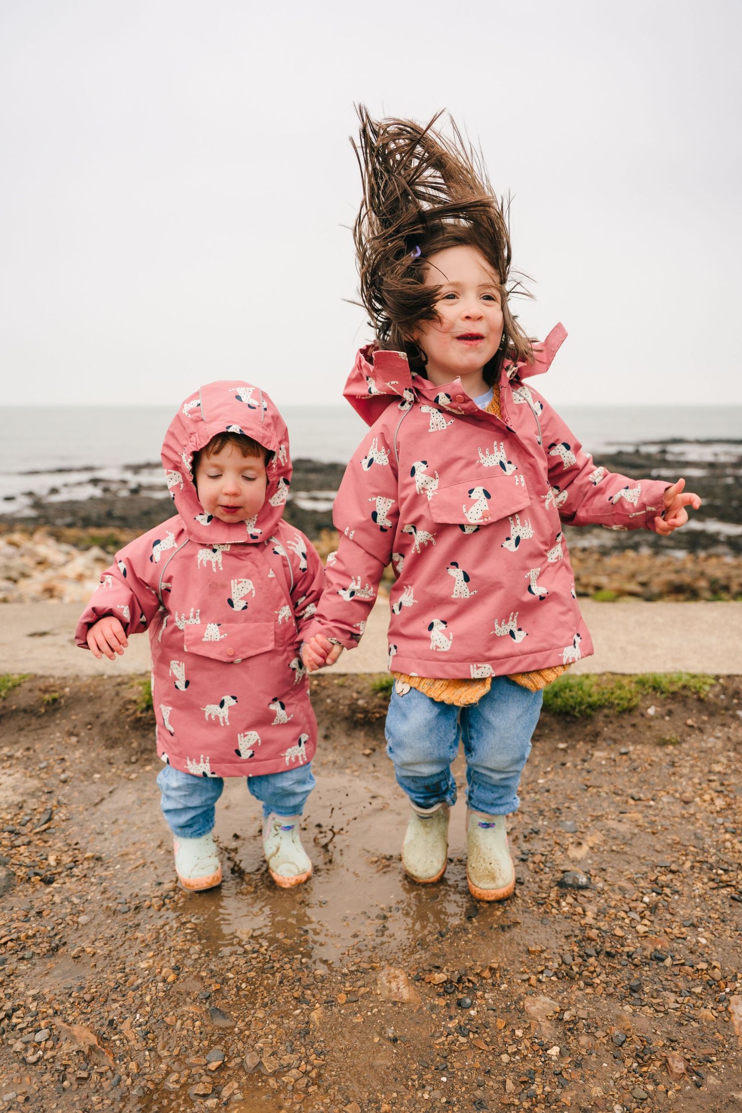 Two children in matching pink penguin raincoats jump together on a muddy coastal path. PHOTOGRAPHER Dublin
