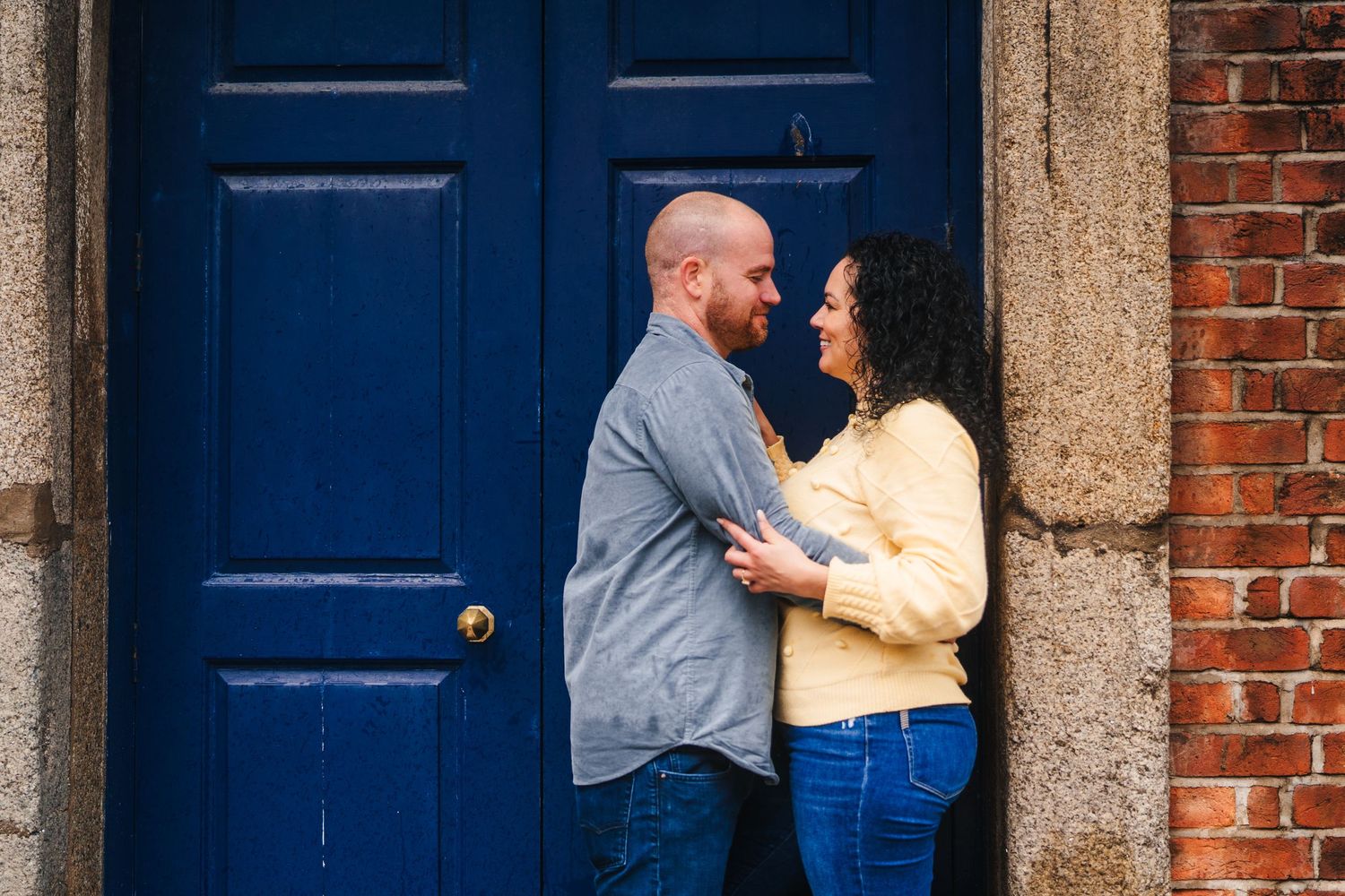 A couple stands affectionately in front of a blue door against a brick wall backdrop. Proposal Dublin Photographer