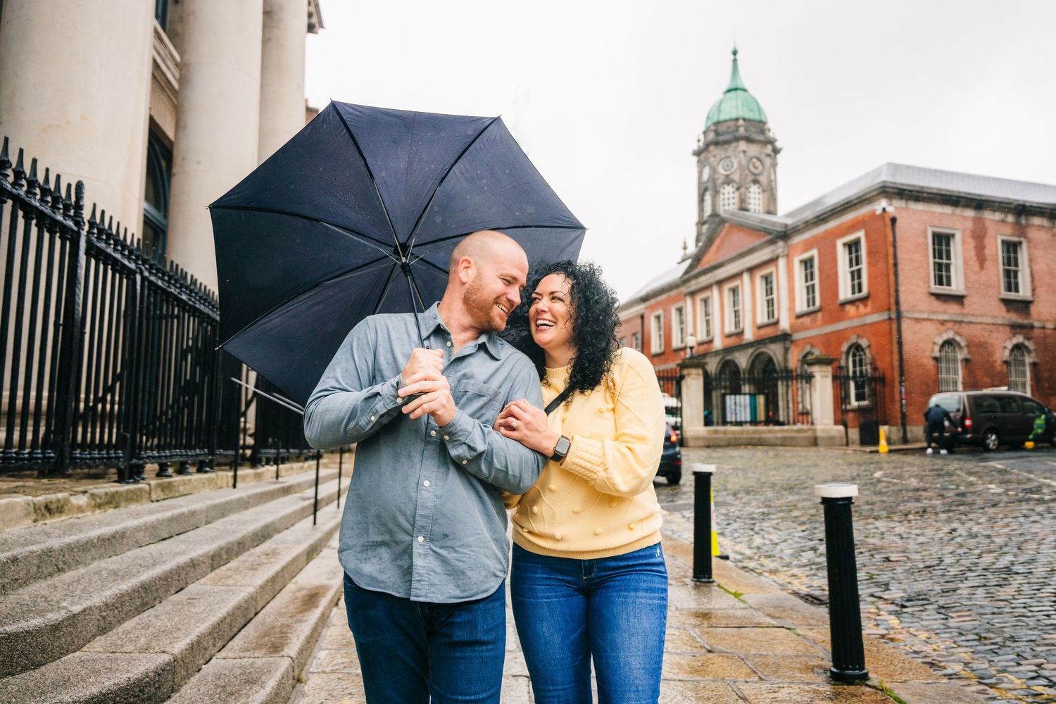Romantic couple walking with umbrella along historic brick buildings and iron railings on rainy day. Proposal Dublin Photographer PHOTOGRAPH