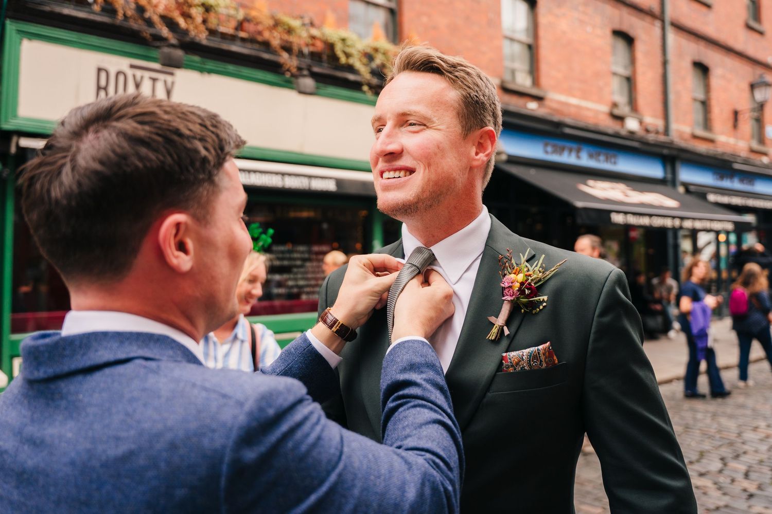 Wedding party members in navy suits share a laugh outside historic brick buildings on a sunny day. Same sex wedding PHOTOGRAPHER Dublin