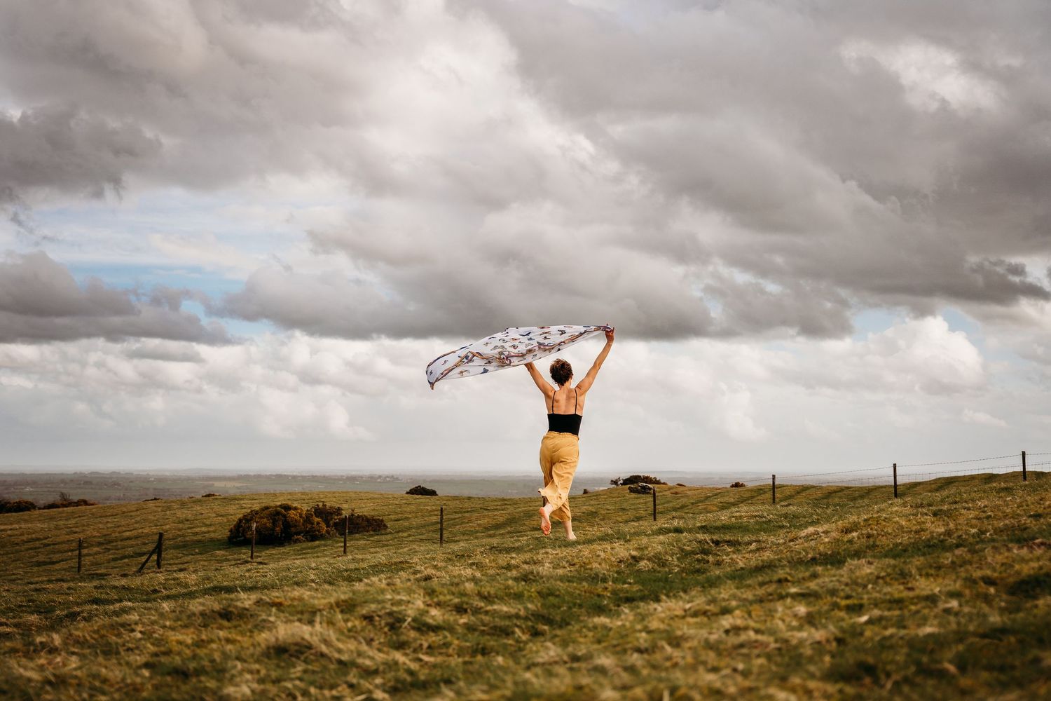 A person in yellow stands on a hilltop with a flowing scarf under dramatic storm clouds in an open landscape. Portrait PHOTOGRAPHER Dublin