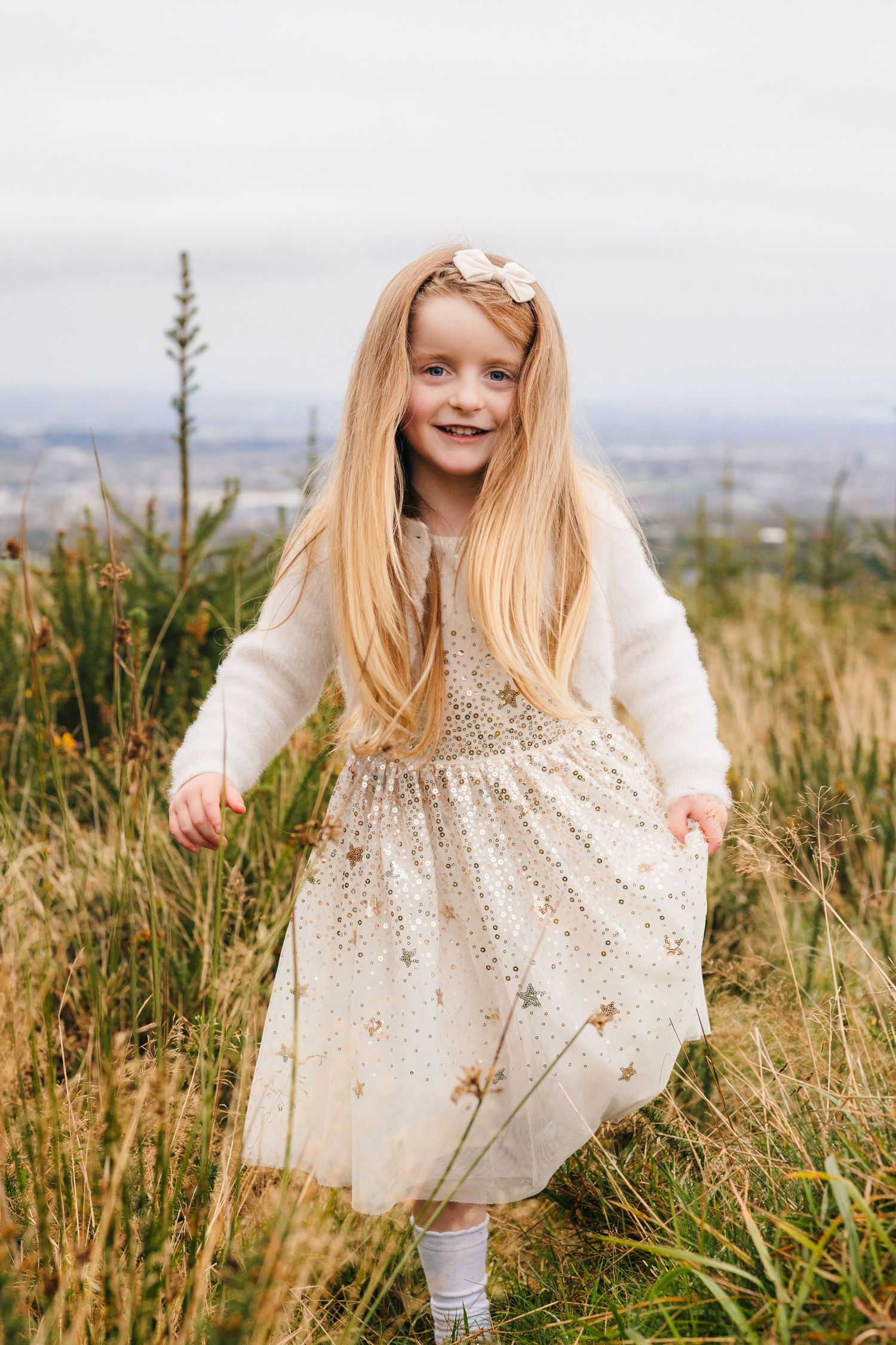 Family Photographer Dublin Ireland A young child in a white lace dress twirls in a field of tall grass against an overcast sky.