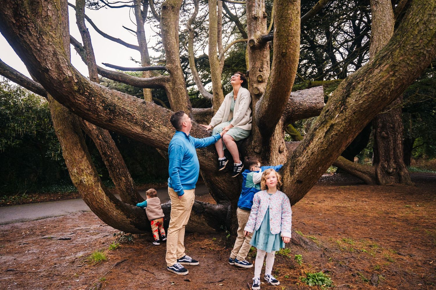 Family members stand and embrace beneath large twisted tree branches in a wooded outdoor setting during golden hour. Family photographer Dub