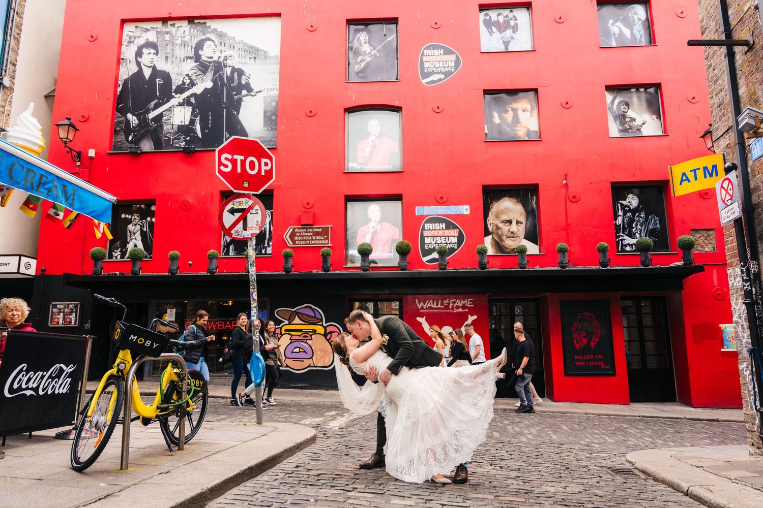 Wedding couple posing in front of red wall with vintage photographs and a parked bicycle. PHOTOGRAPHER Dublin Ireland Wedding