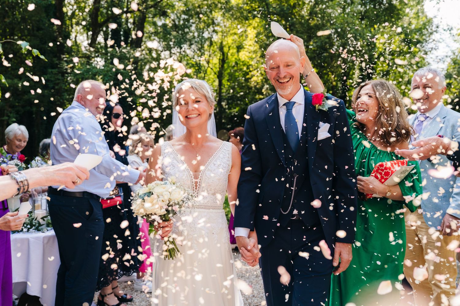 Bride and groom walk through confetti shower surrounded by smiling wedding guests outdoors.
