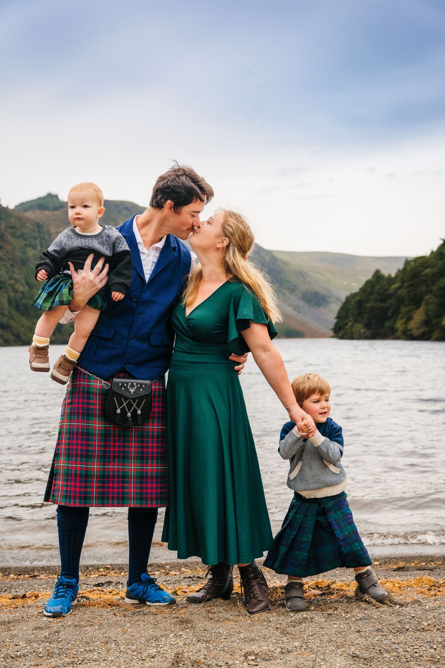 Family in traditional Scottish attire poses together by a scenic mountain lake.Family photogrAPHER DUBLIN