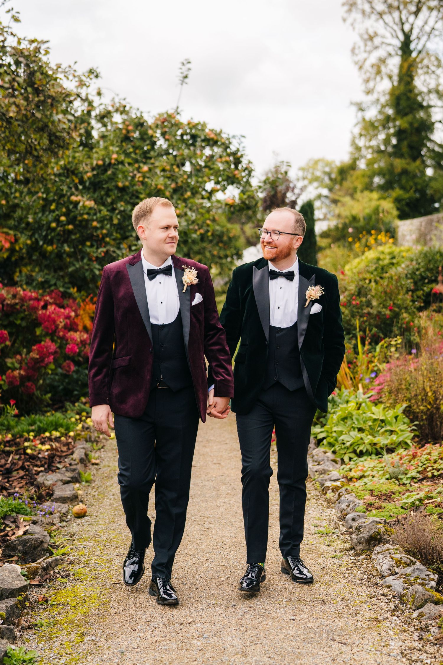 Two people in burgundy and black tuxedos walk together on a garden path surrounded by autumn foliage. LGBTQ Ashley Park Wedding Photographer