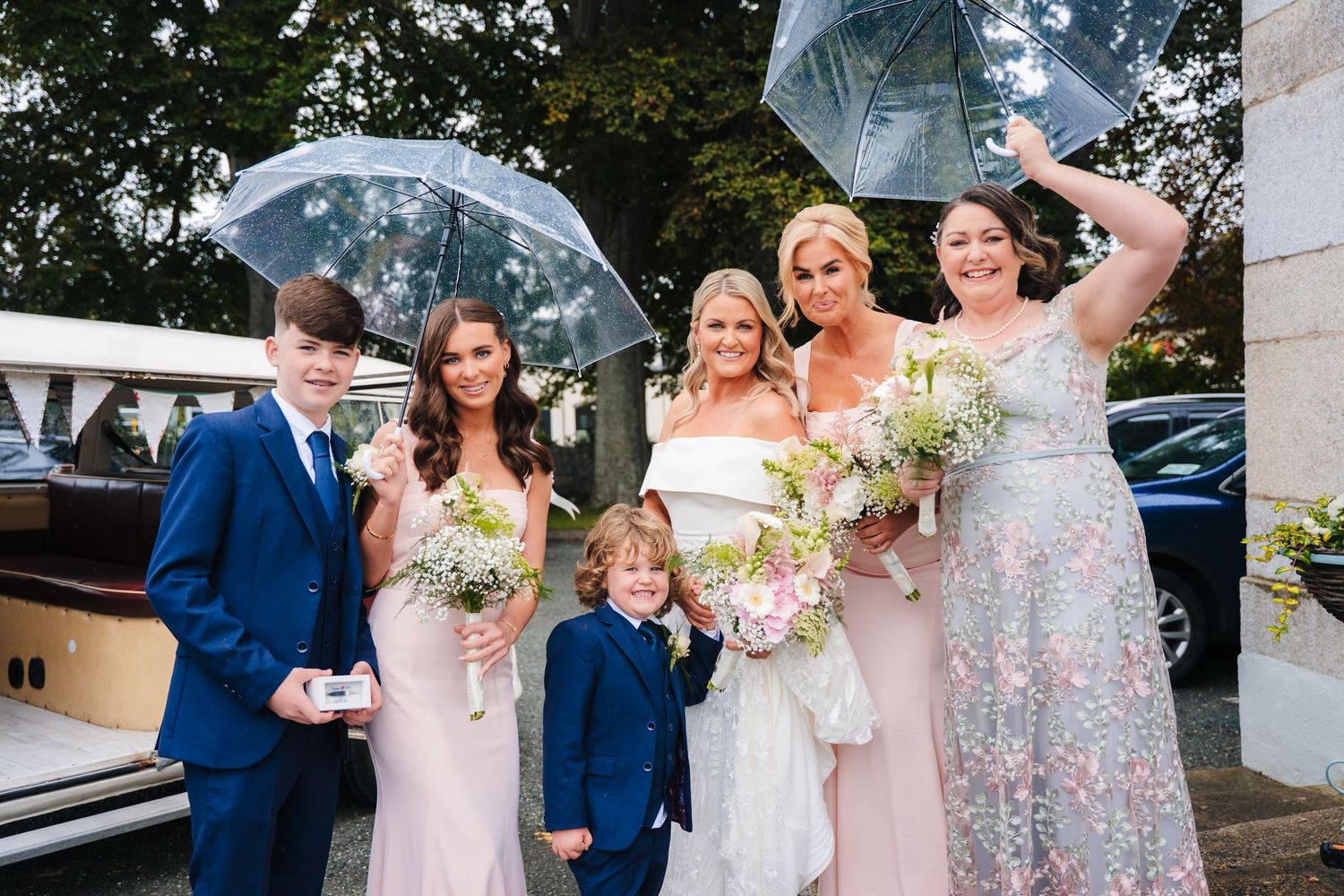 Wedding party poses joyfully with clear umbrellas on a rainy day. The Dean Dublin Wedding Photographer