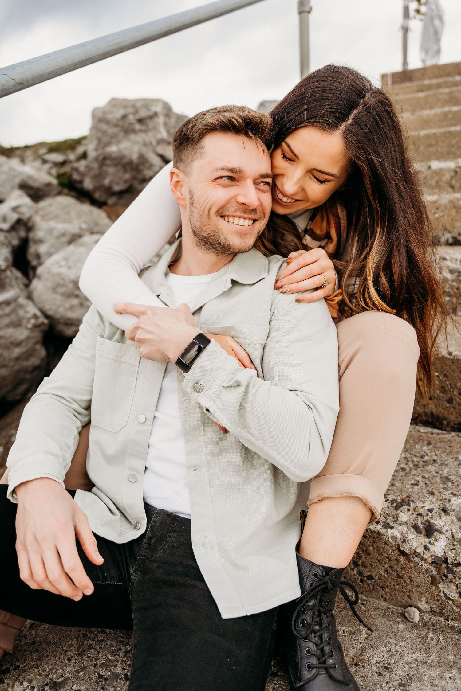 A couple embraces and laughs together while sitting on coastal rocks in muted tones. PHOTOGRAPHER Dublin ireland