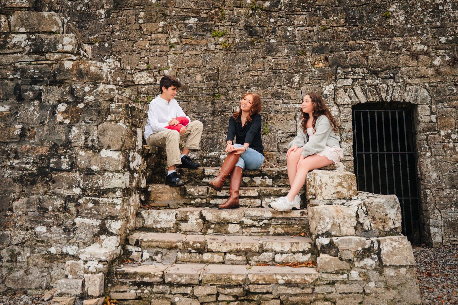 People sitting casually along an old stone wall at a historic site. BEctive abbey family photographer dublin
