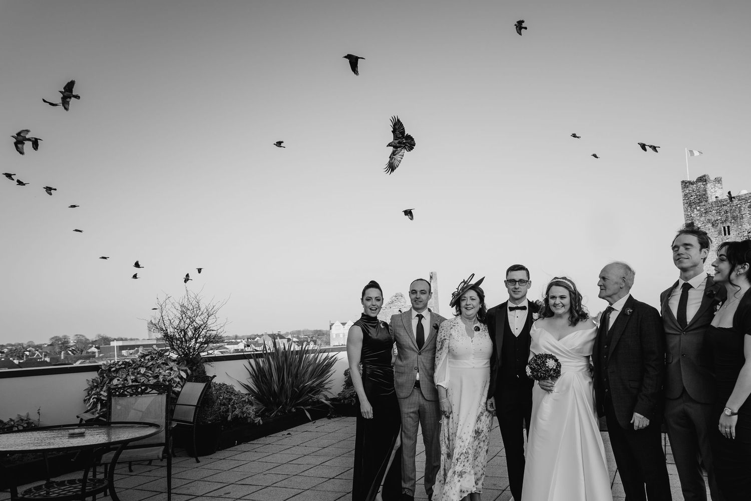 Birds flying across a grey sky above a wedding gathering in black and white.  Wedding PHOTOGRAPHER Trim Castle