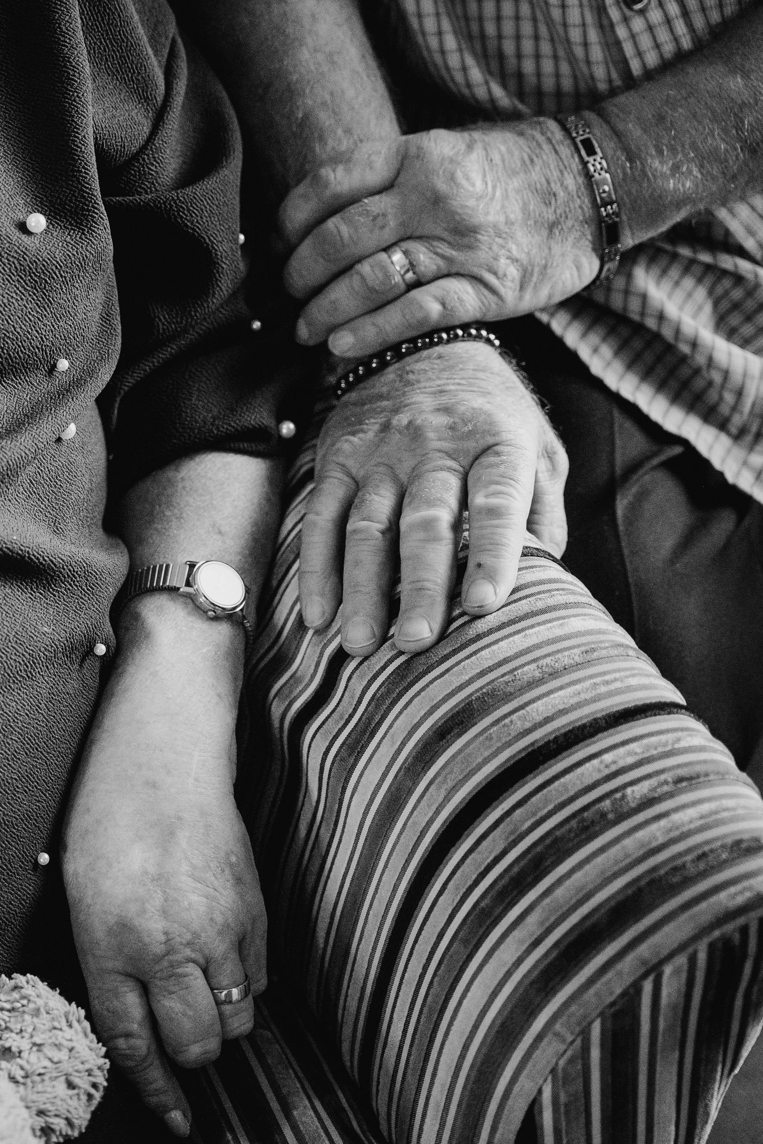 Close-up black and white photograph of aged hands with watches resting on striped fabric. Wedding PHOTOGRAPHER Trim