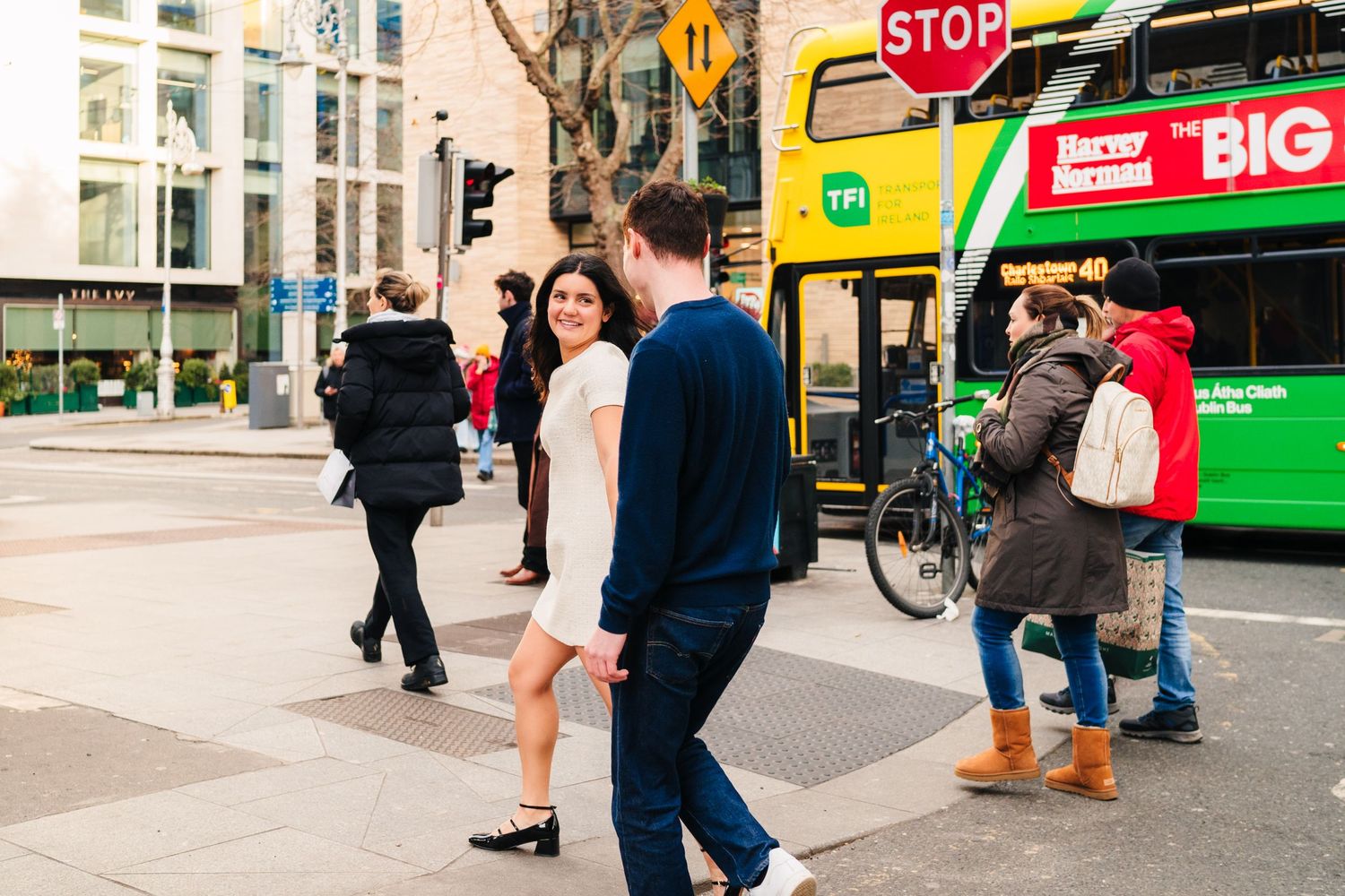 People walk past shops and a city bus on a busy urban street with storefronts and stop signs visible. Surprise proposal dublin photography