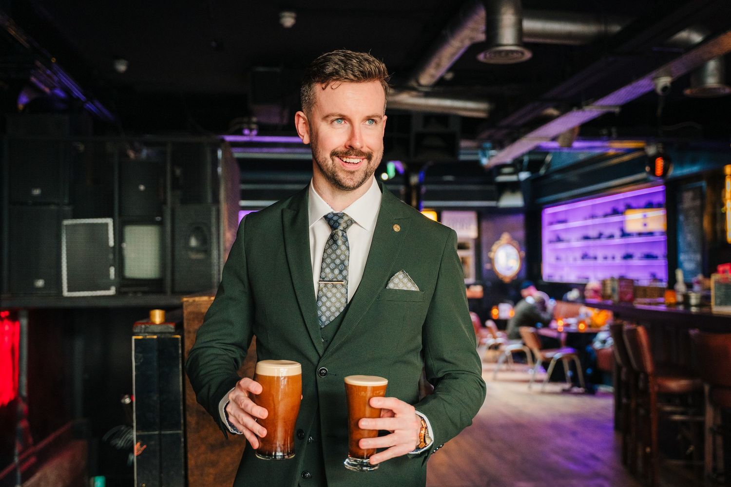 Man in green blazer holding two craft beer glasses in dimly lit pub setting. City Hall Dublin wedding photogrAPher
