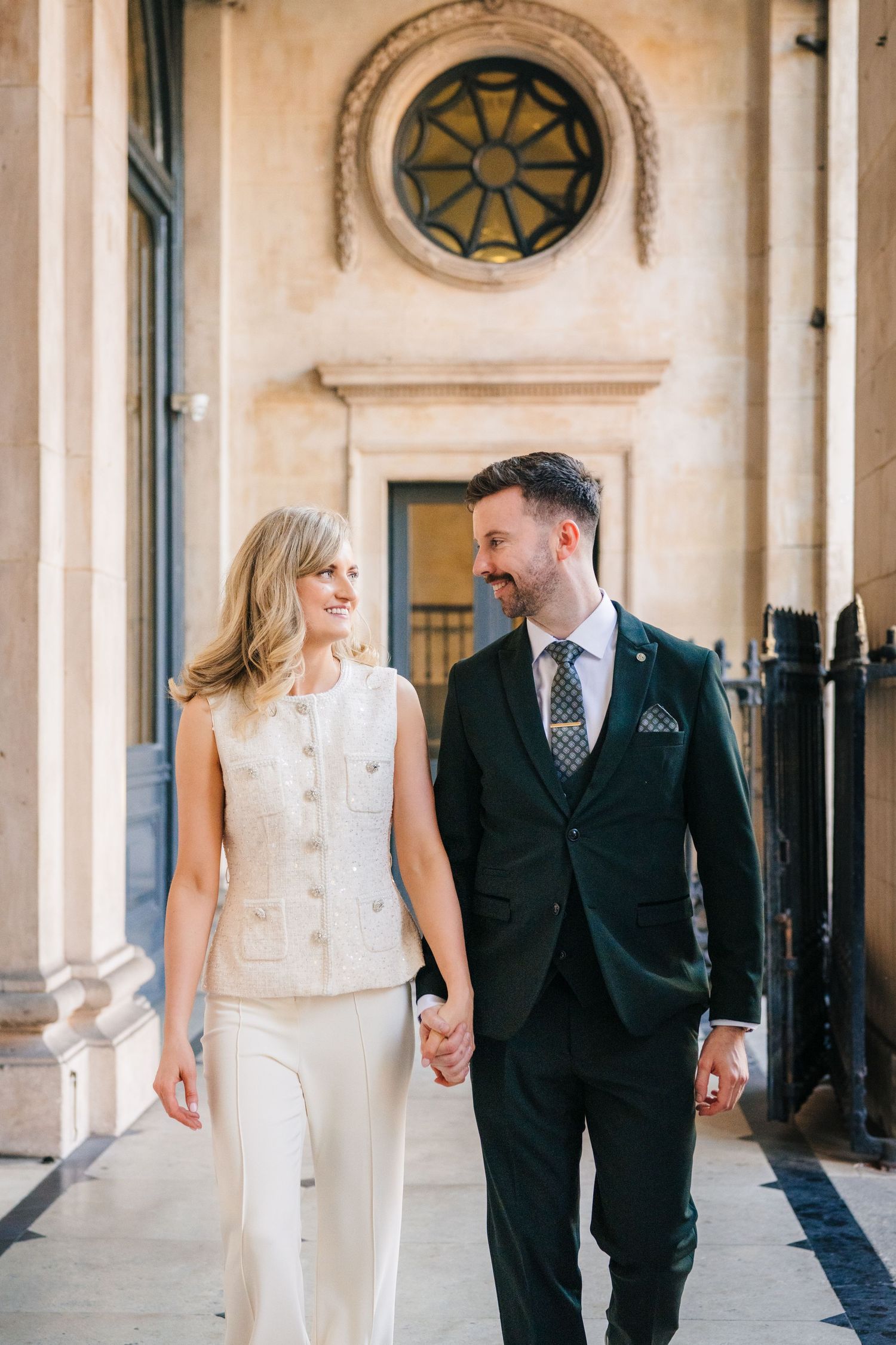 Couple holding hands walks under architectural archway with circular window detail. City Hall Dublin wedding photogrAPher