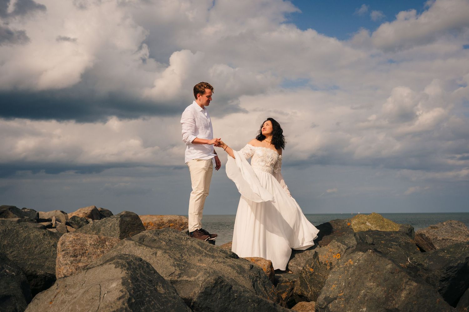 A couple in white outfits shares a romantic moment on rocky coastal cliffs against a dramatic cloudy sky. Wedding PHOTOGRAPHER Dublin