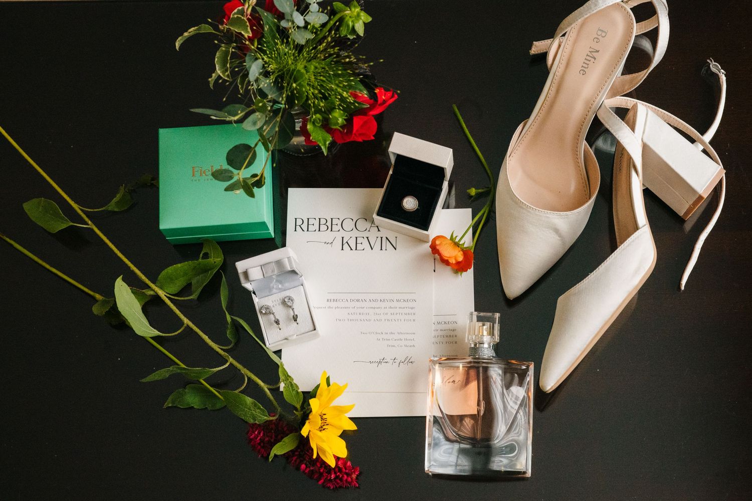 Wedding details flatlay with white heels, invitation, perfume bottle, green box, and red and yellow flowers on dark background. Wedding Phot