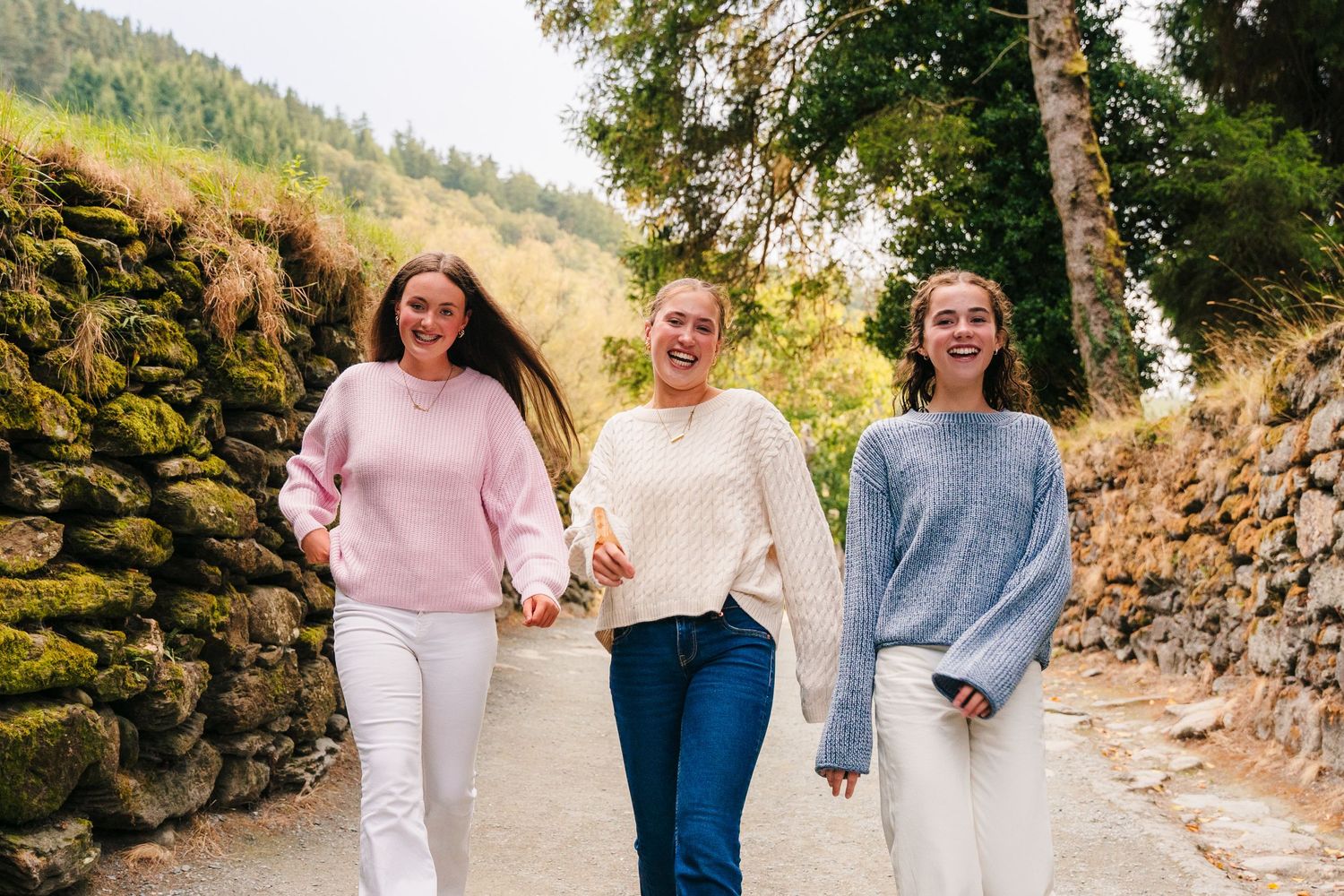 Three friends in cozy sweaters walk together on a path beside a stone wall on a sunny autumn day. Family photographer Ireland Dublin