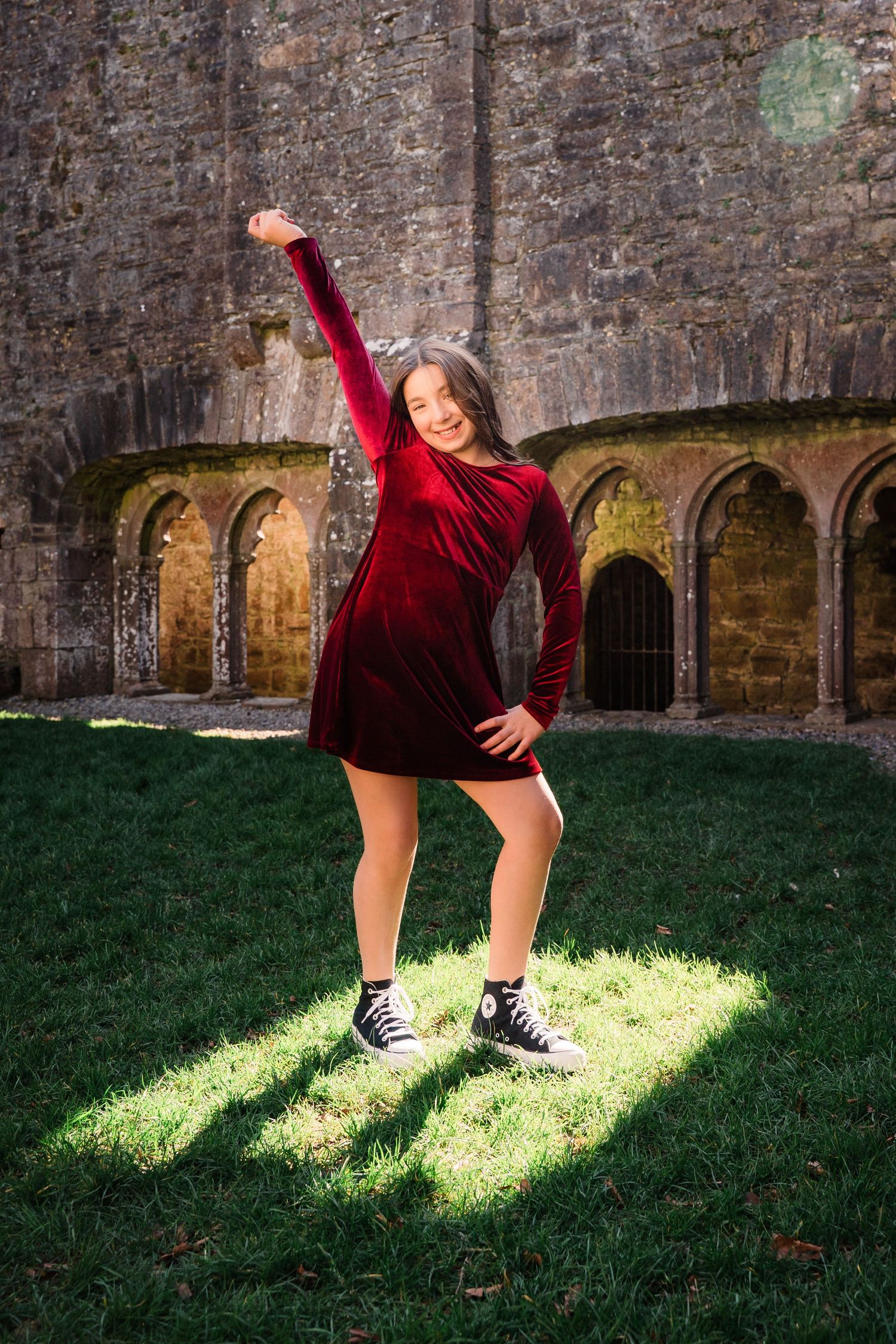 A joyful pose in a red velvet dress and sneakers against medieval castle ruins and arches. Family PHOTOGRAPHER Dublin
