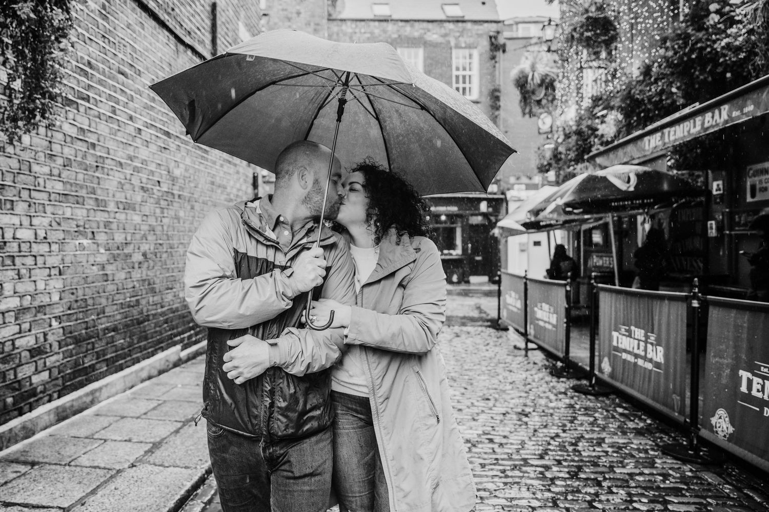 A romantic couple holding umbrellas shares an intimate moment in the rain on a cobblestone street. Proposal Dublin Photographer