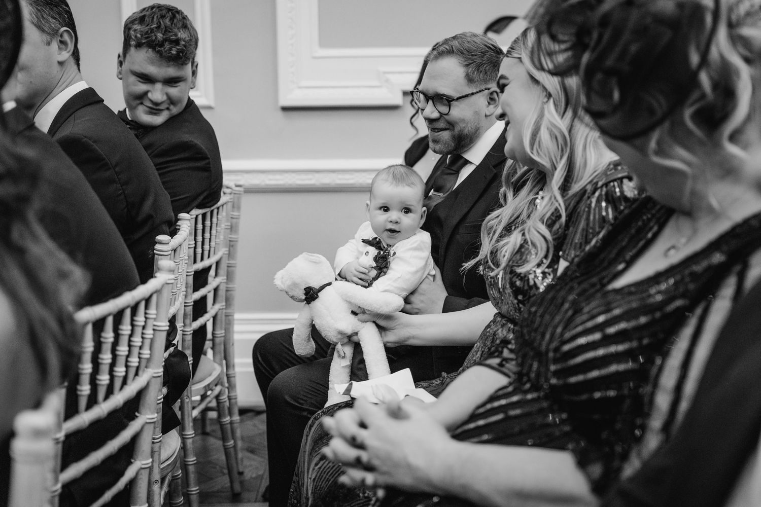A black and white photo of guests at a formal event with a baby being held in the crowd. Stephens green club dublin Wedding photographer