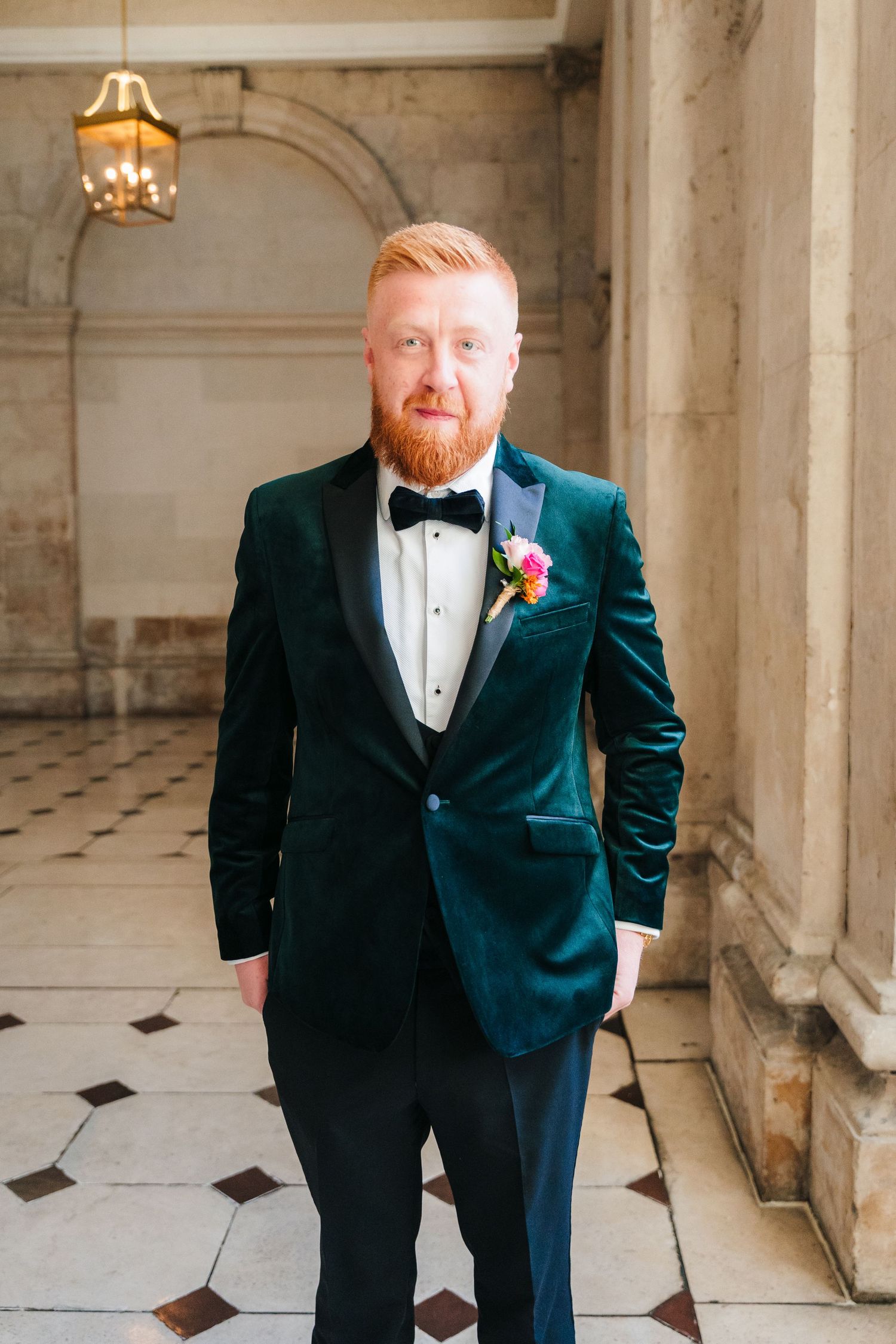 A groom in an emerald green velvet tuxedo poses in an elegant columned hallway with marble floors. Wedding photographer dublin